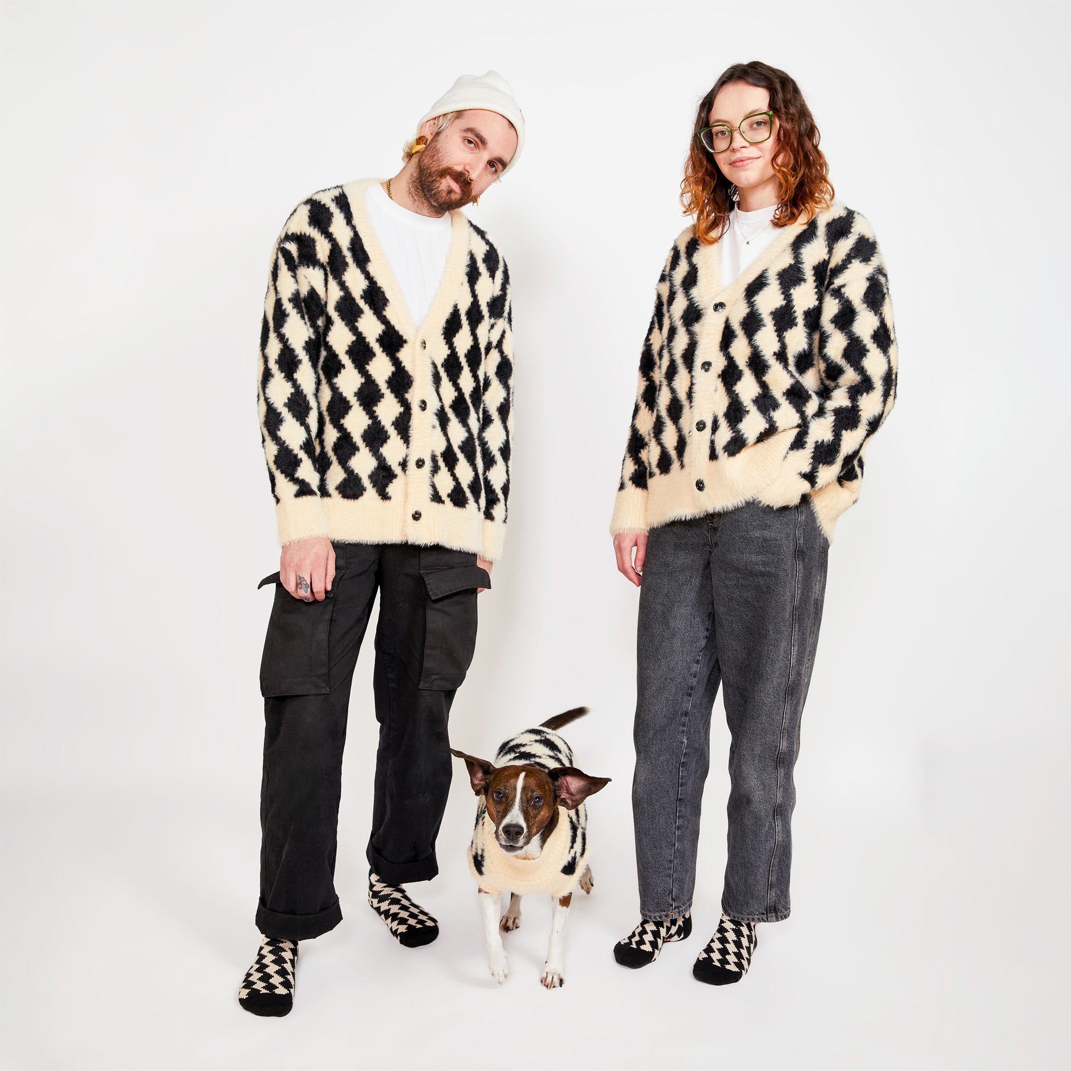 Two people standing with a brown dog between them. All three are wearing matching black and cream diamond-patterned knitwear, with the people also wearing matching socks.