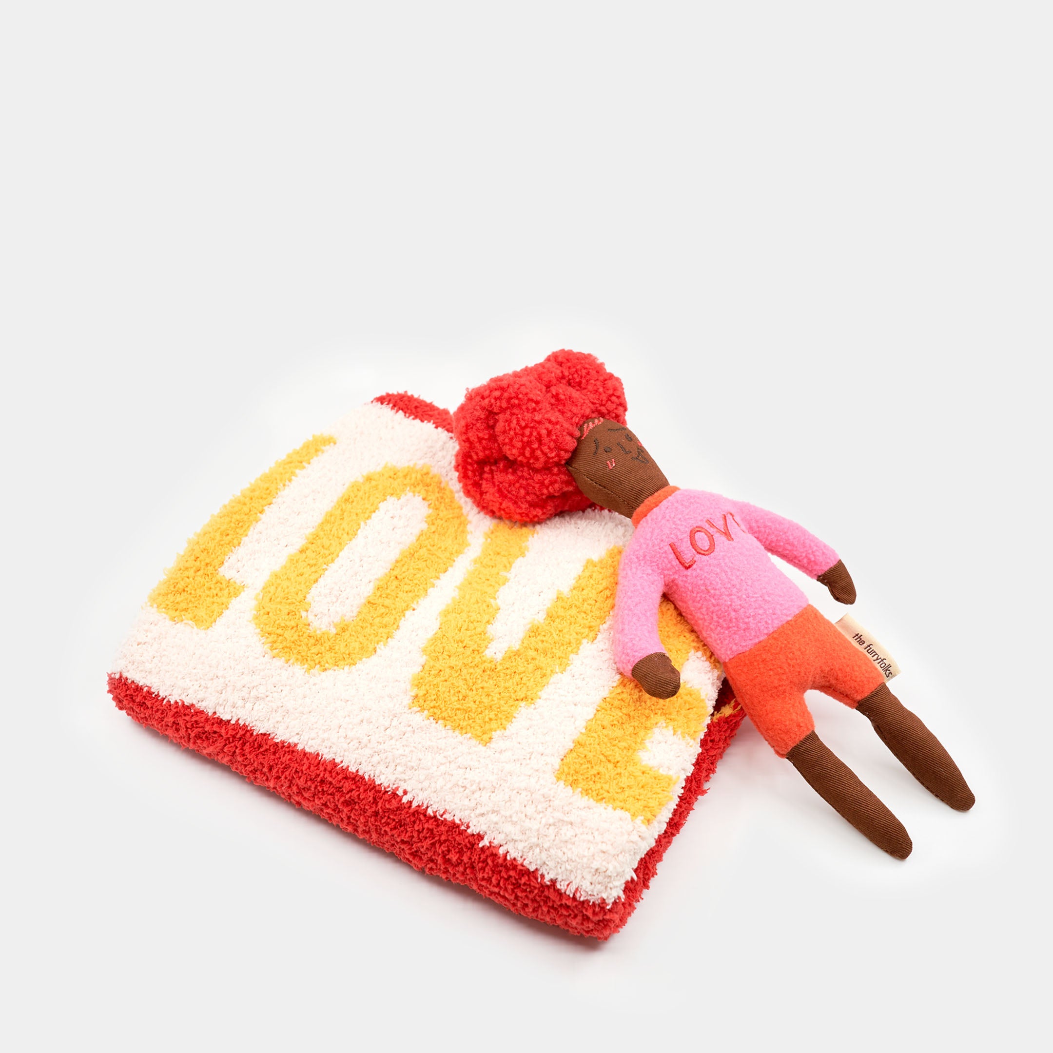 The photograph shows a red-haired, pink-shirted doll-shaped nosework toy resting on a plush dog bed with "LOVE" written in bold yellow letters. The toy's red pom-pom hair and pink "LOVE" shirt echo the bed's design, creating a thematic pairing that's visually appealing. This setup is likely designed to provide a comfortable and engaging environment for a dog, encouraging play and relaxation. The white background emphasizes the toy and bed's vibrant colors and the heartfelt message.