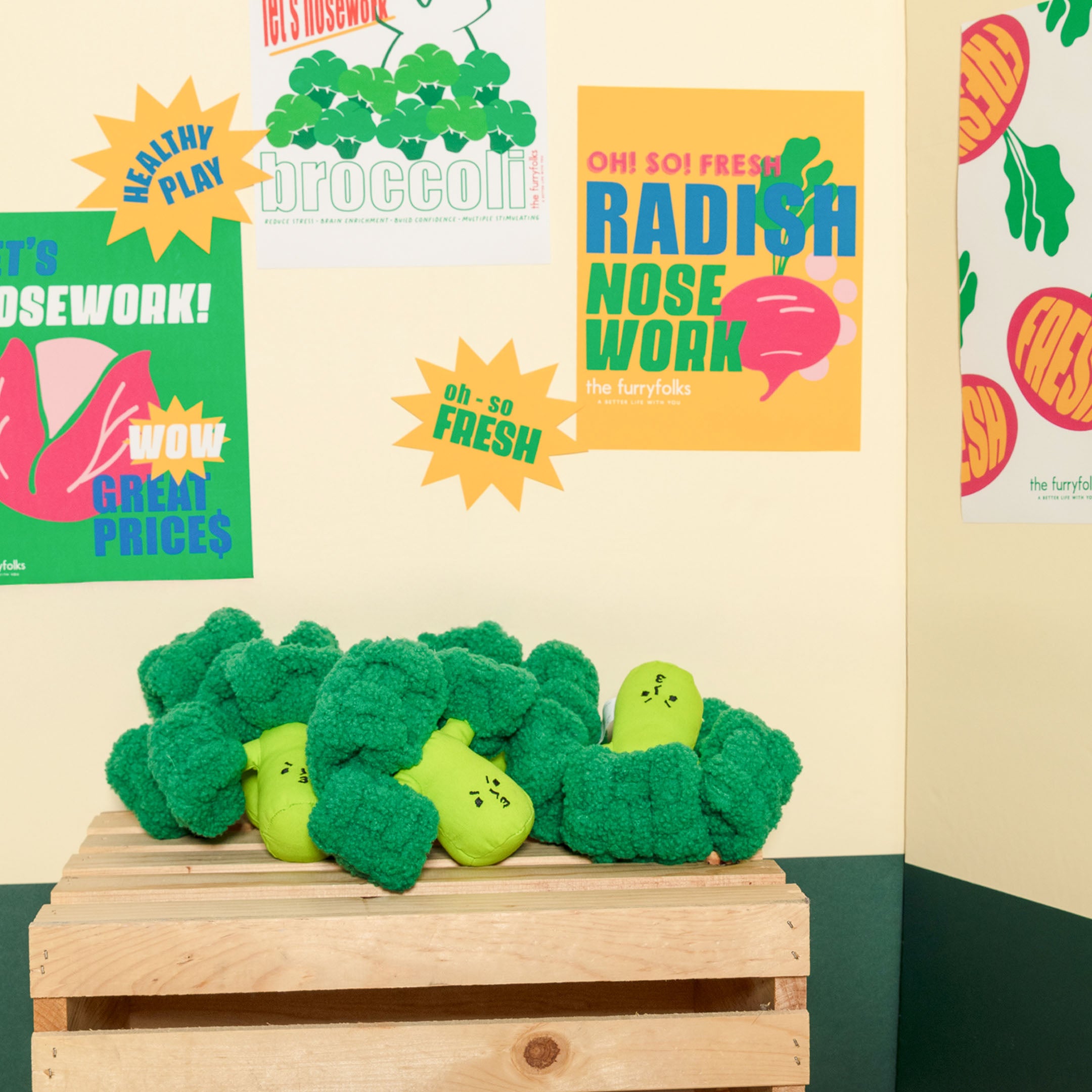 Green plush toy resembling broccoli on a wooden crate with colorful posters in the background.