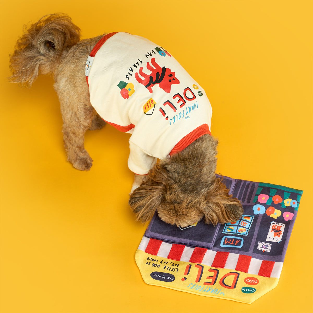 Small brown dog wearing a cream deli-print shirt sniffs the purple door flap of the Deli Nosework mat on a bright yellow floor.