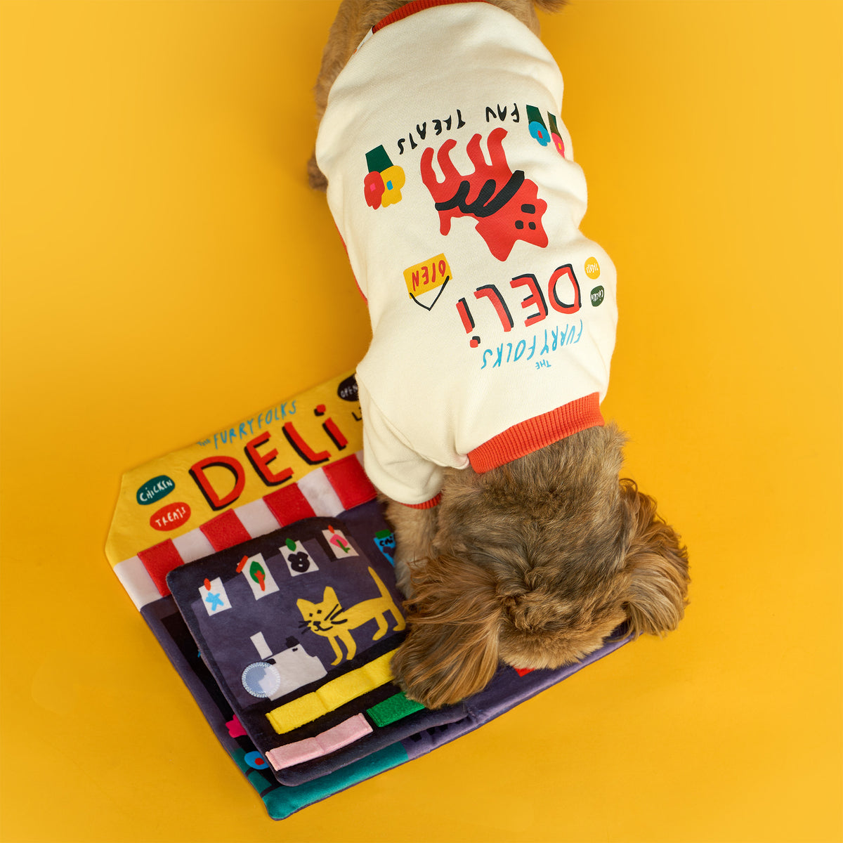 Top view of a small brown dog in a cream deli-print shirt sniffing the open Deli Nosework mat, with colorful fabric loops on a bright yellow floor.