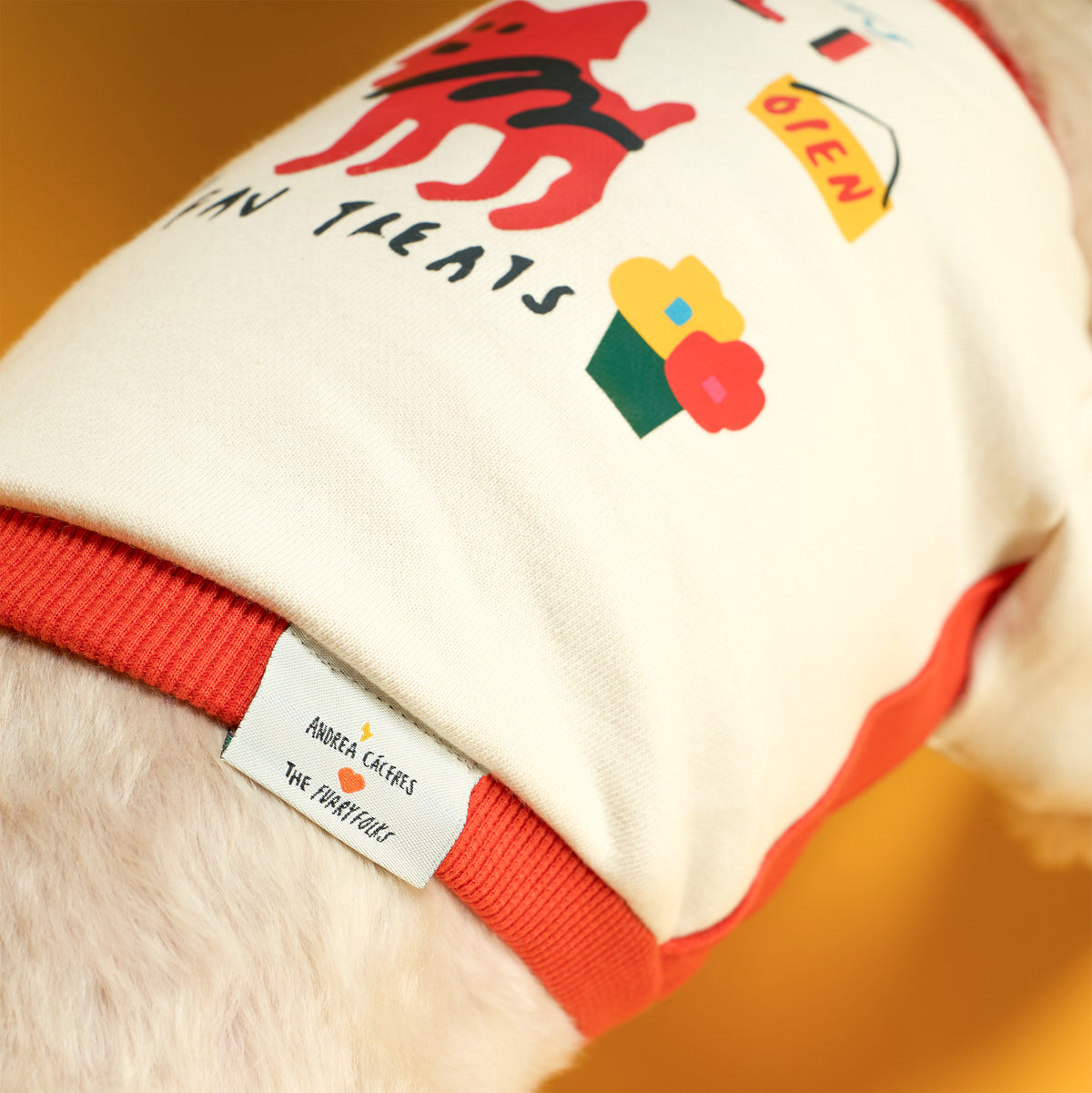Close-up of a cream dog shirt with orange ribbed trim, colorful dog and flower graphics, and a woven “Andrea Cáceres ♥ the furryfolks” label, worn by a light-furred dog.