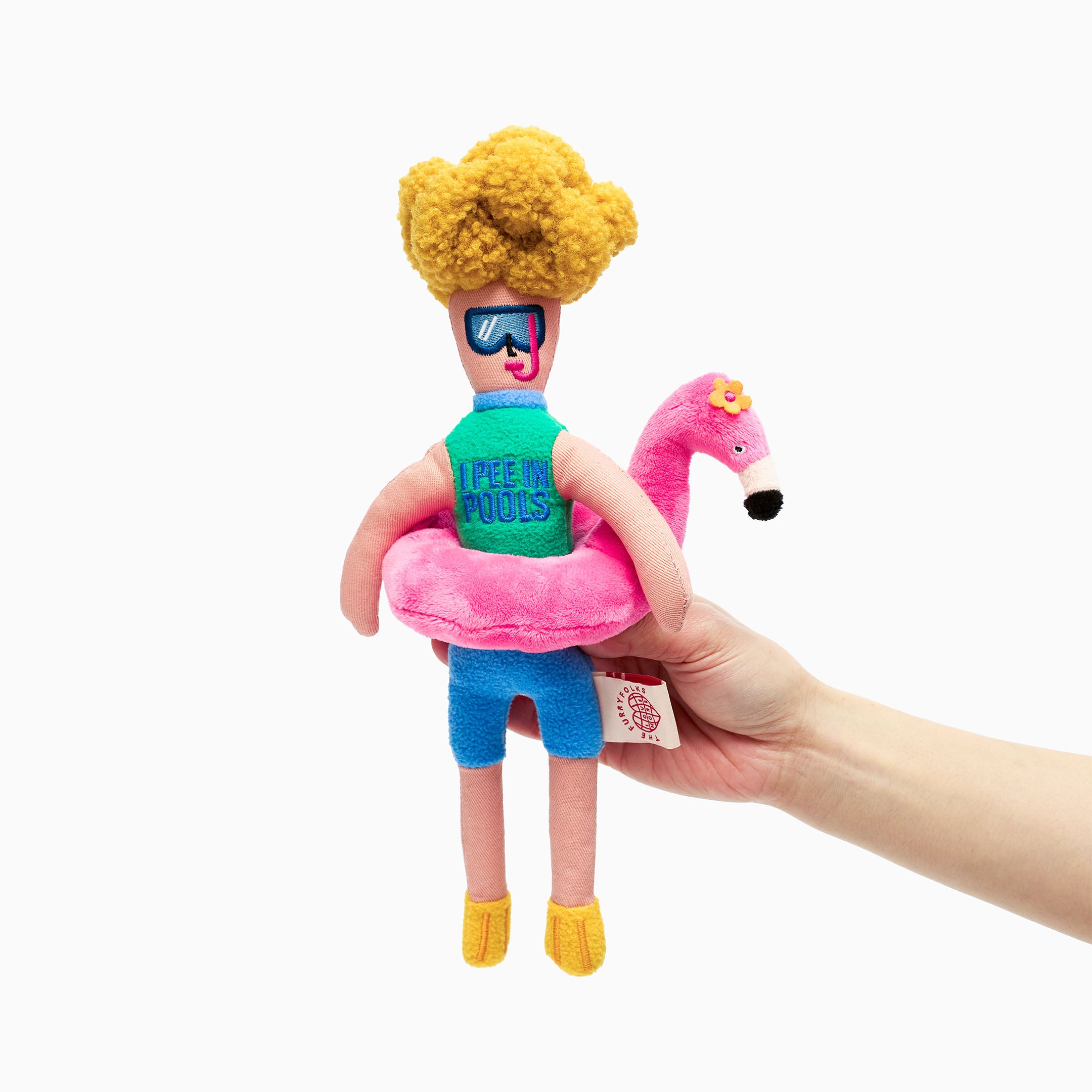 A plush dog toy shaped like a sun-loving human with yellow curly hair, sunglasses, a green tank that says “I PEE IN POOLS,” wearing a pink flamingo float and yellow flippers, being held upright by a hand.