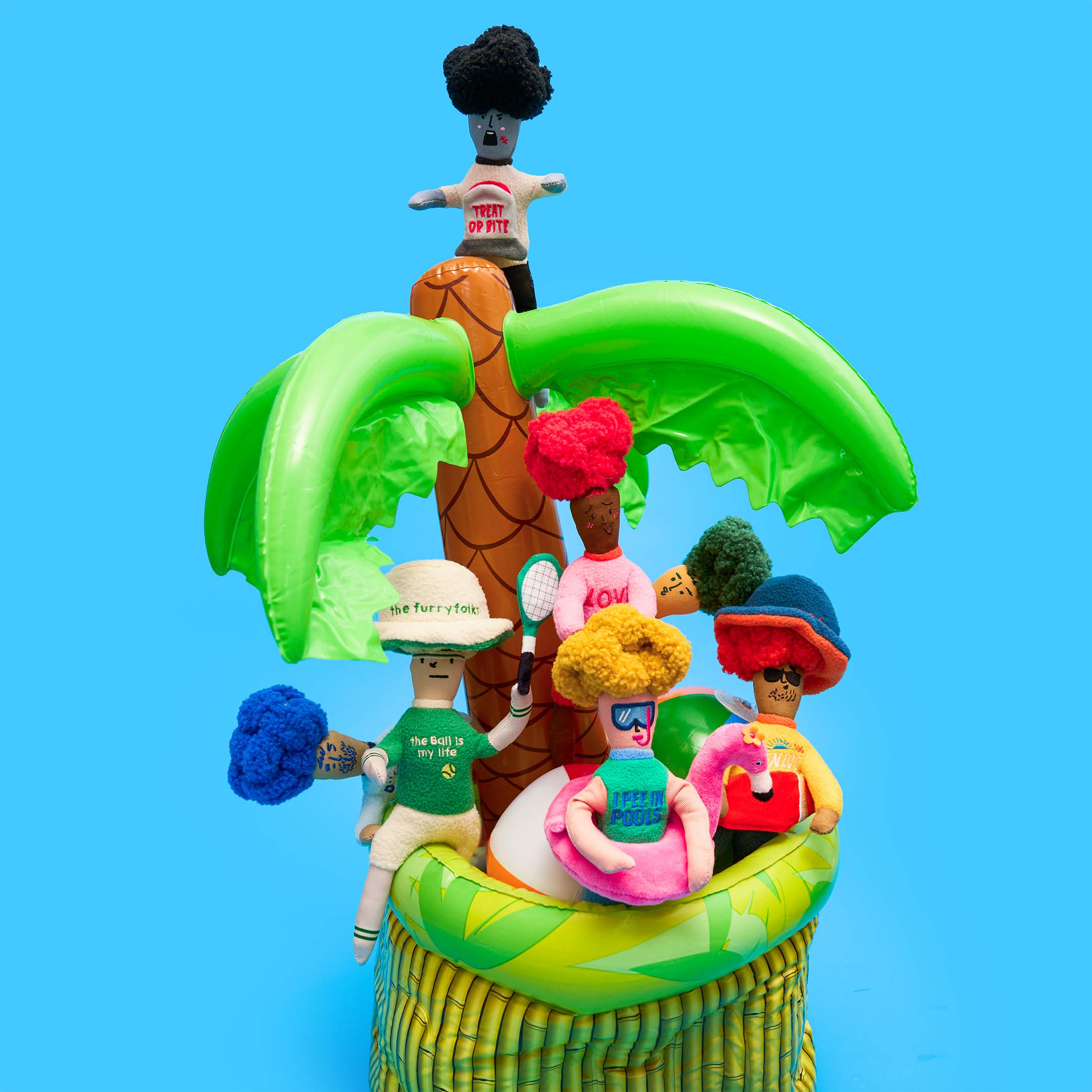 Several plush Hooman toys from the furryfolks, including Flamingo Hooman, are arranged around an inflatable palm tree and tropical-themed setup on a blue background.