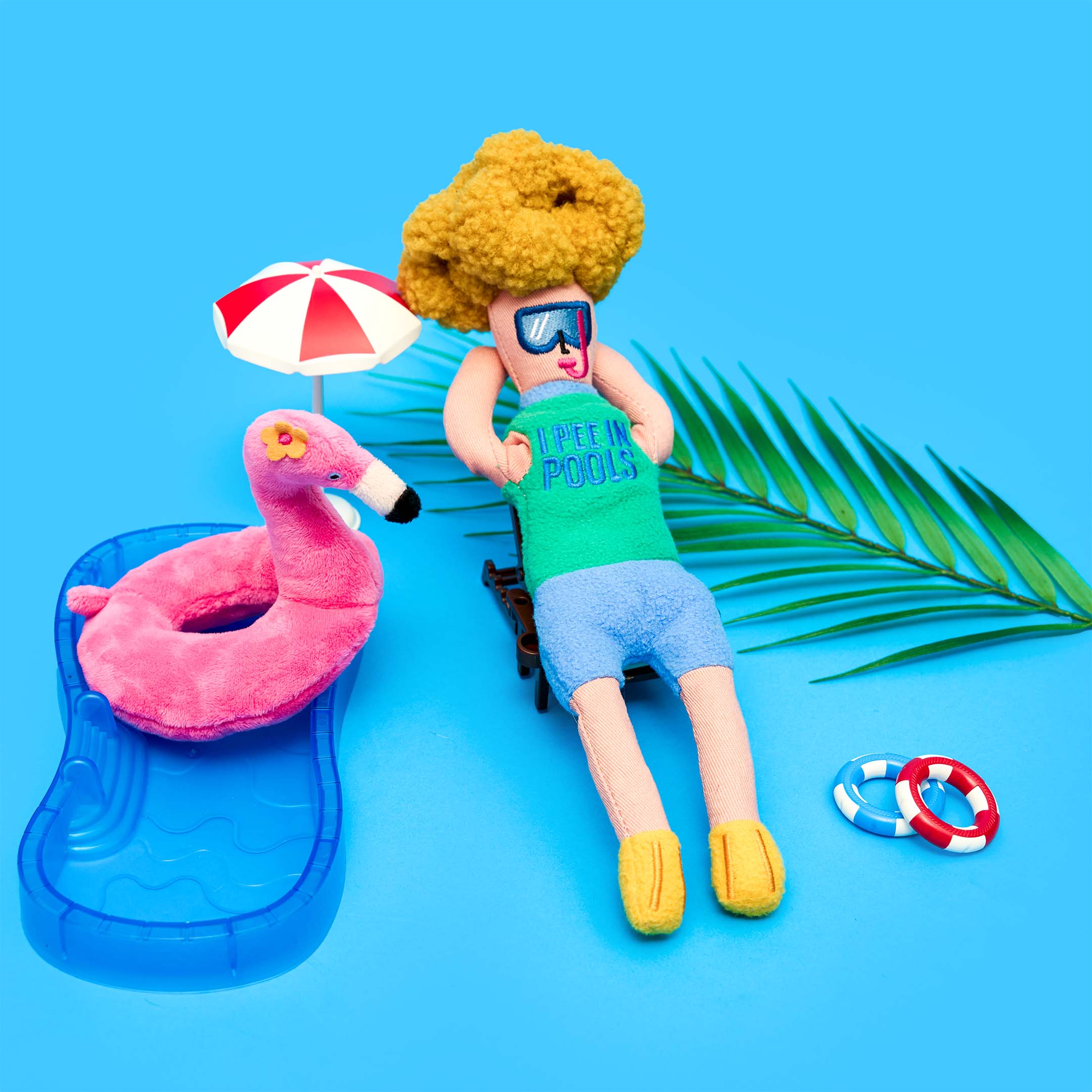 The Flamingo Hooman plush toy lounges beside a pink flamingo float toy, next to a mini pool, beach umbrella, palm leaf, and life rings on a bright blue background.