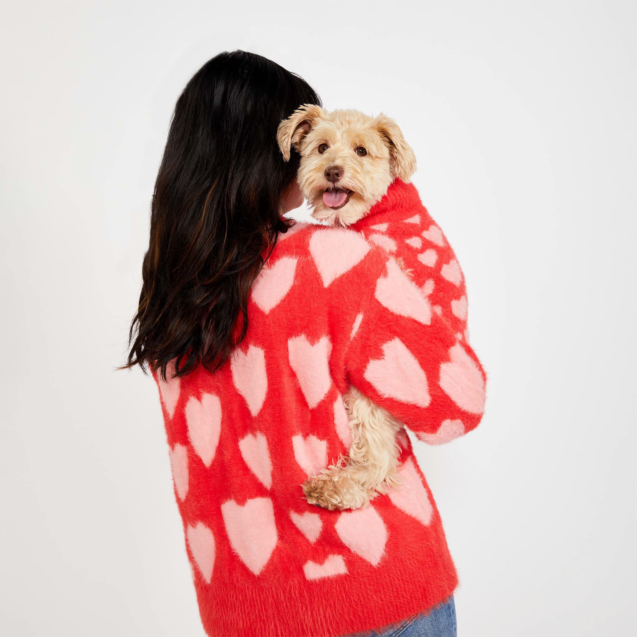 A woman with long dark hair, seen from behind, holds a small beige dog wearing a matching red sweater with pink heart patterns. Both the woman and the dog are wearing the same heart-patterned knitwear.