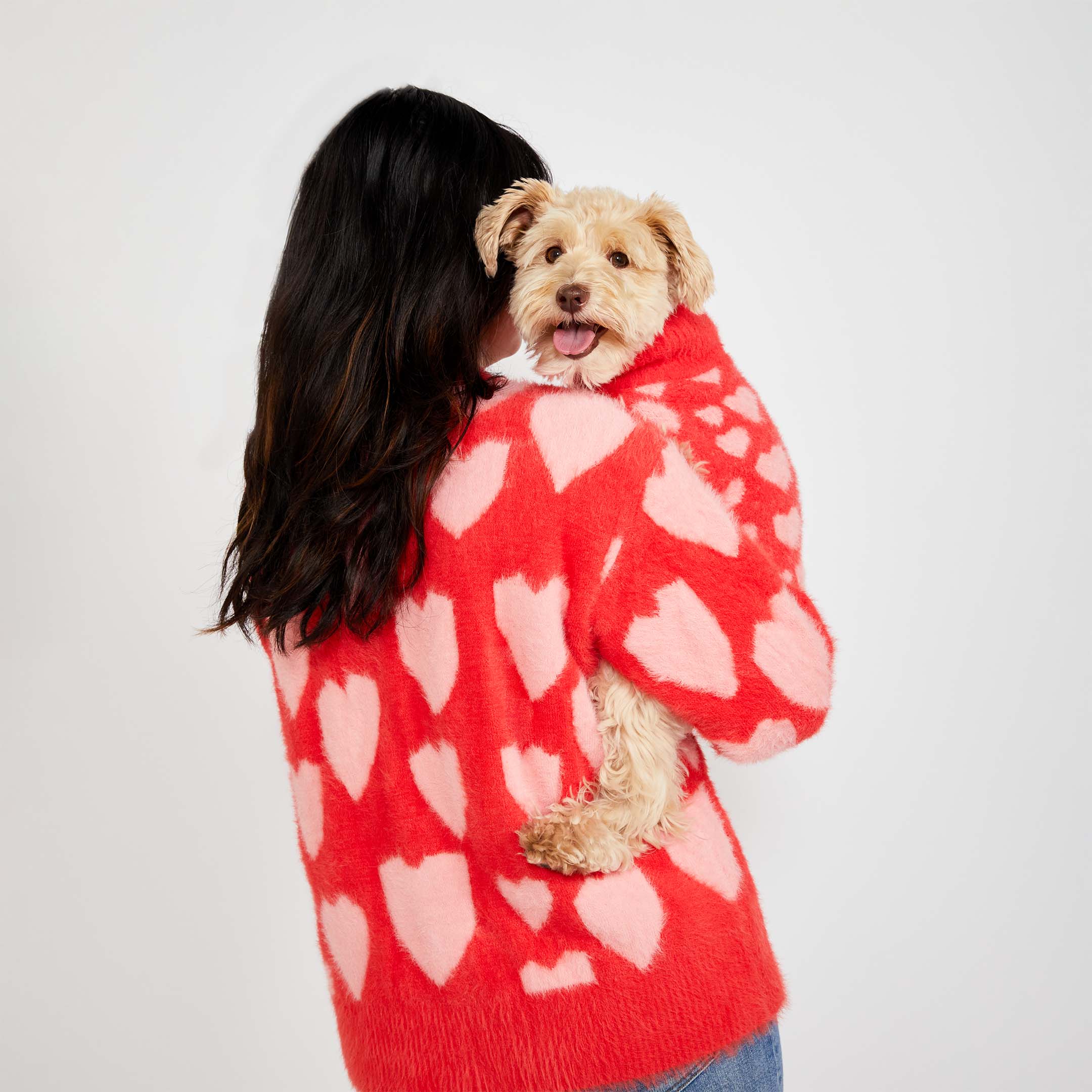 A woman with long dark hair, seen from behind, holds a small beige dog wearing a matching red sweater with pink heart patterns. Both the woman and the dog are wearing the same heart-patterned knitwear.