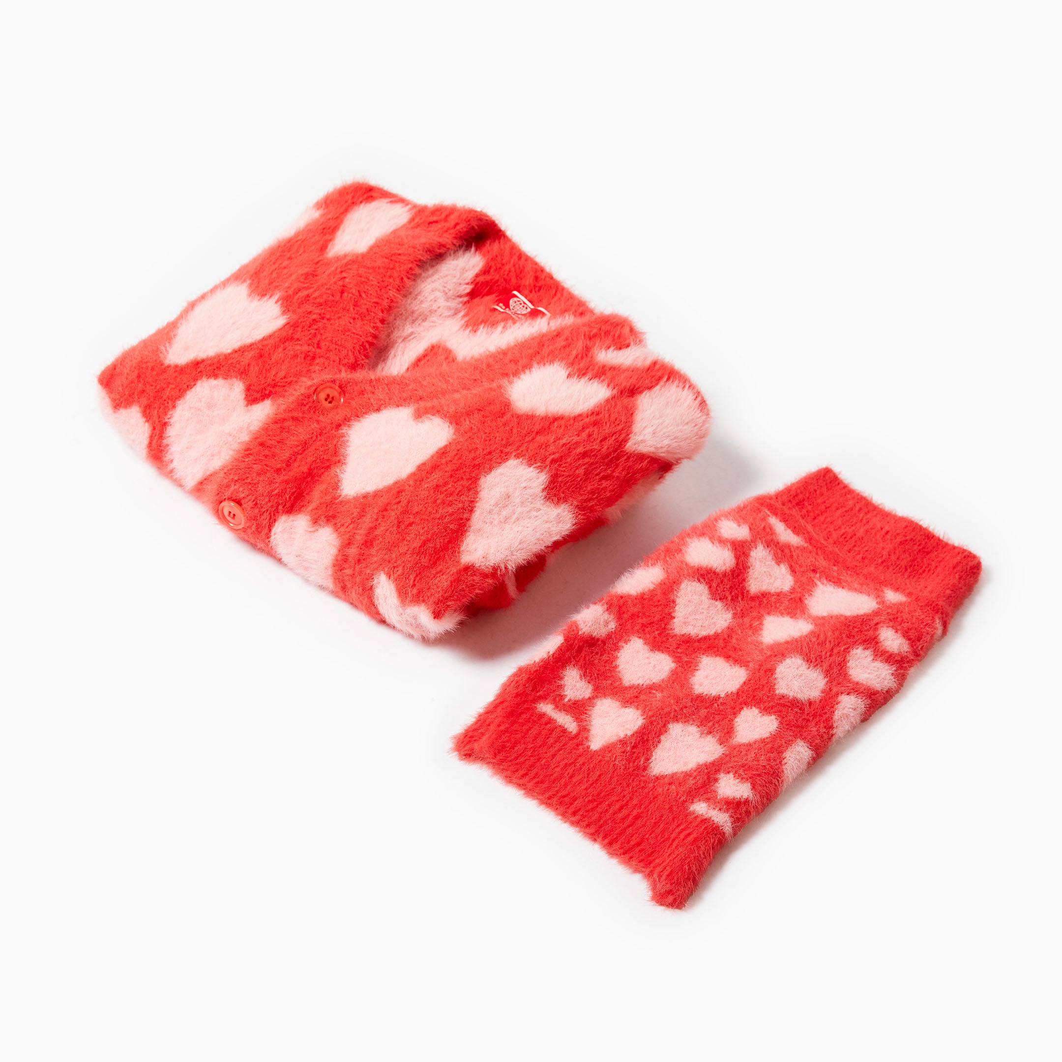 A folded red cardigan with pink hearts is placed beside a matching dog sweater, both showcasing the playful heart design and cozy material.