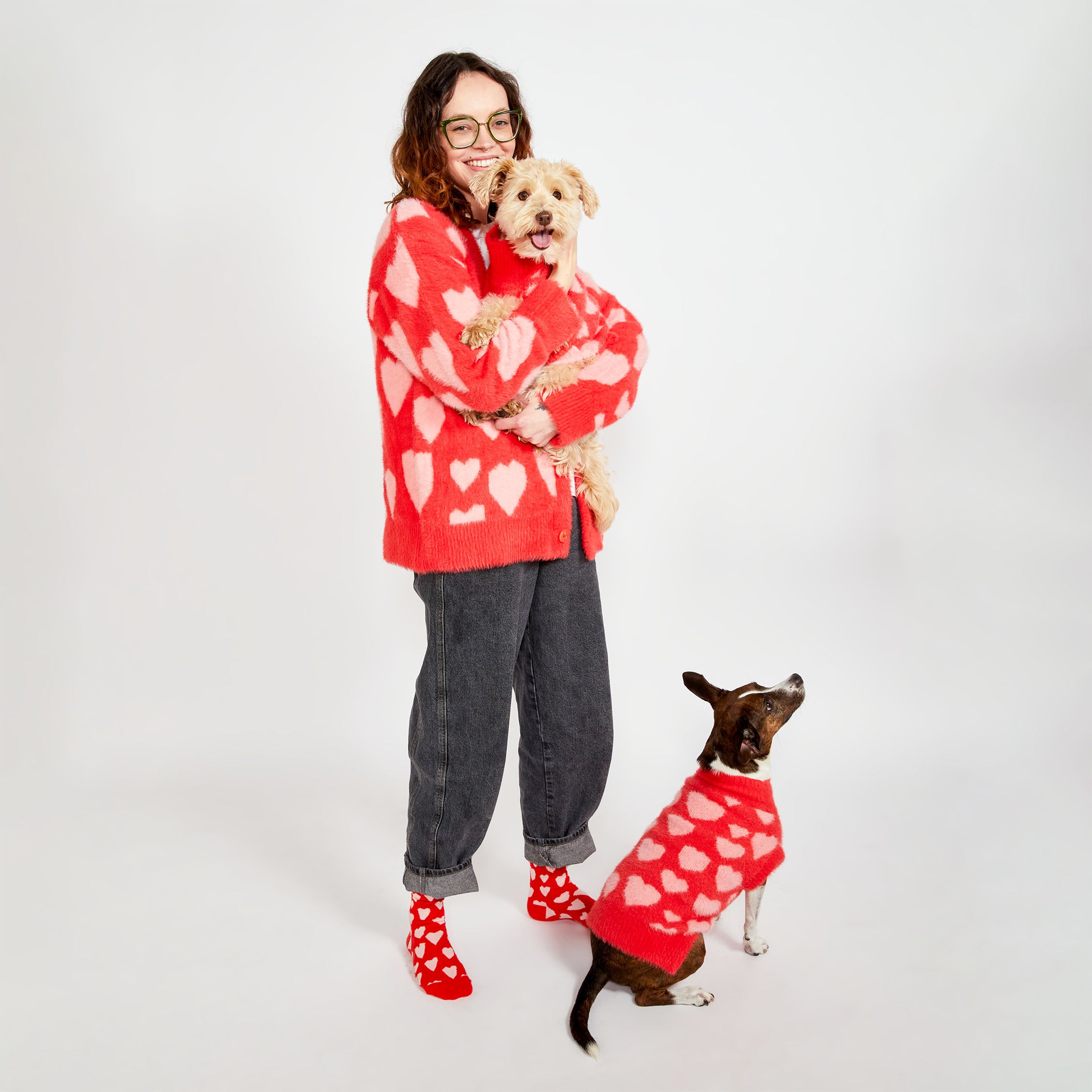 A woman standing and holding a small beige dog while another brown dog sits by her side. Both dogs are wearing red sweaters with pink heart patterns, and the woman is wearing a matching red cardigan with pink hearts and red socks with white hearts.