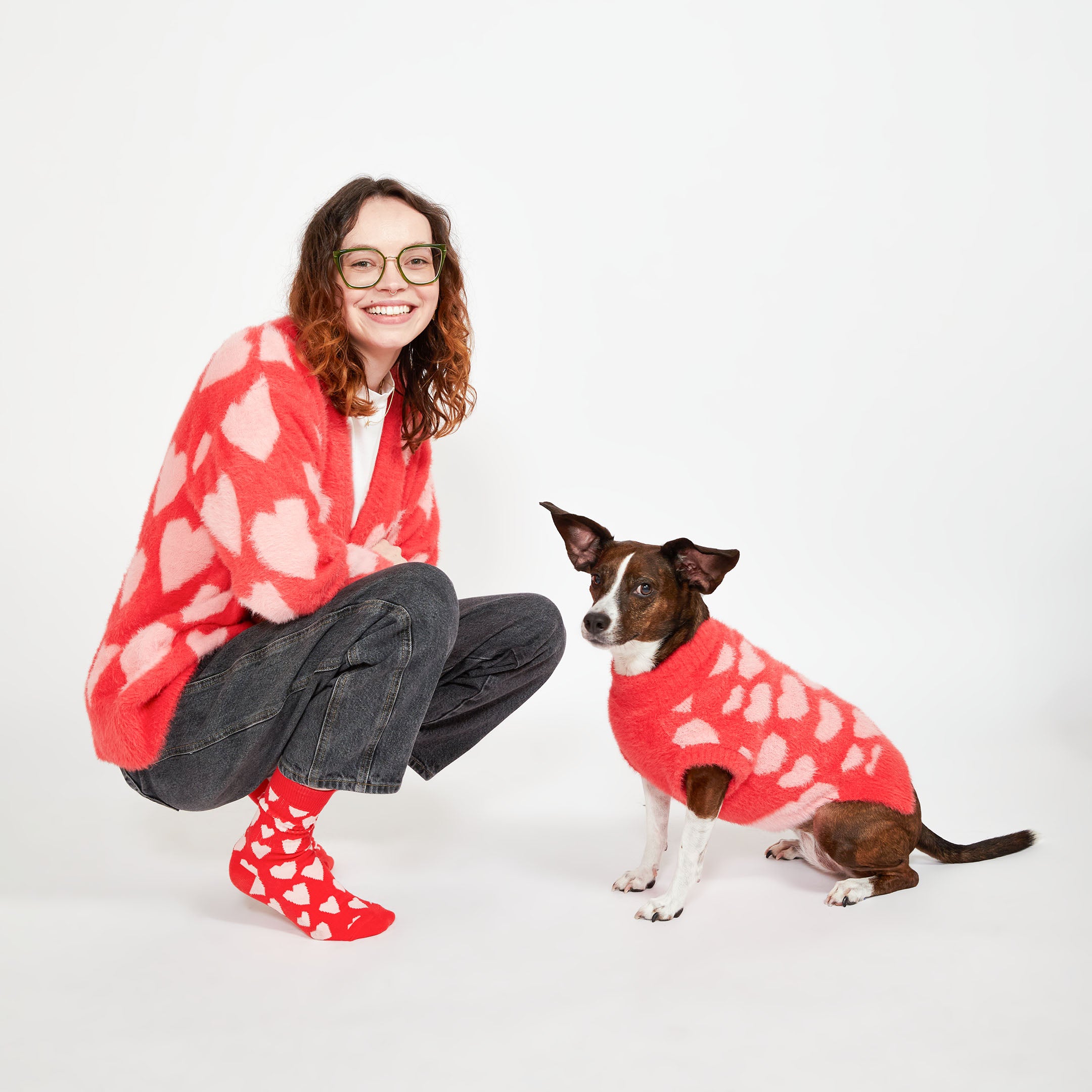 A woman crouching next to a brown dog. Both are wearing matching red sweaters with pink heart patterns. The woman is smiling and wearing red socks with white hearts.