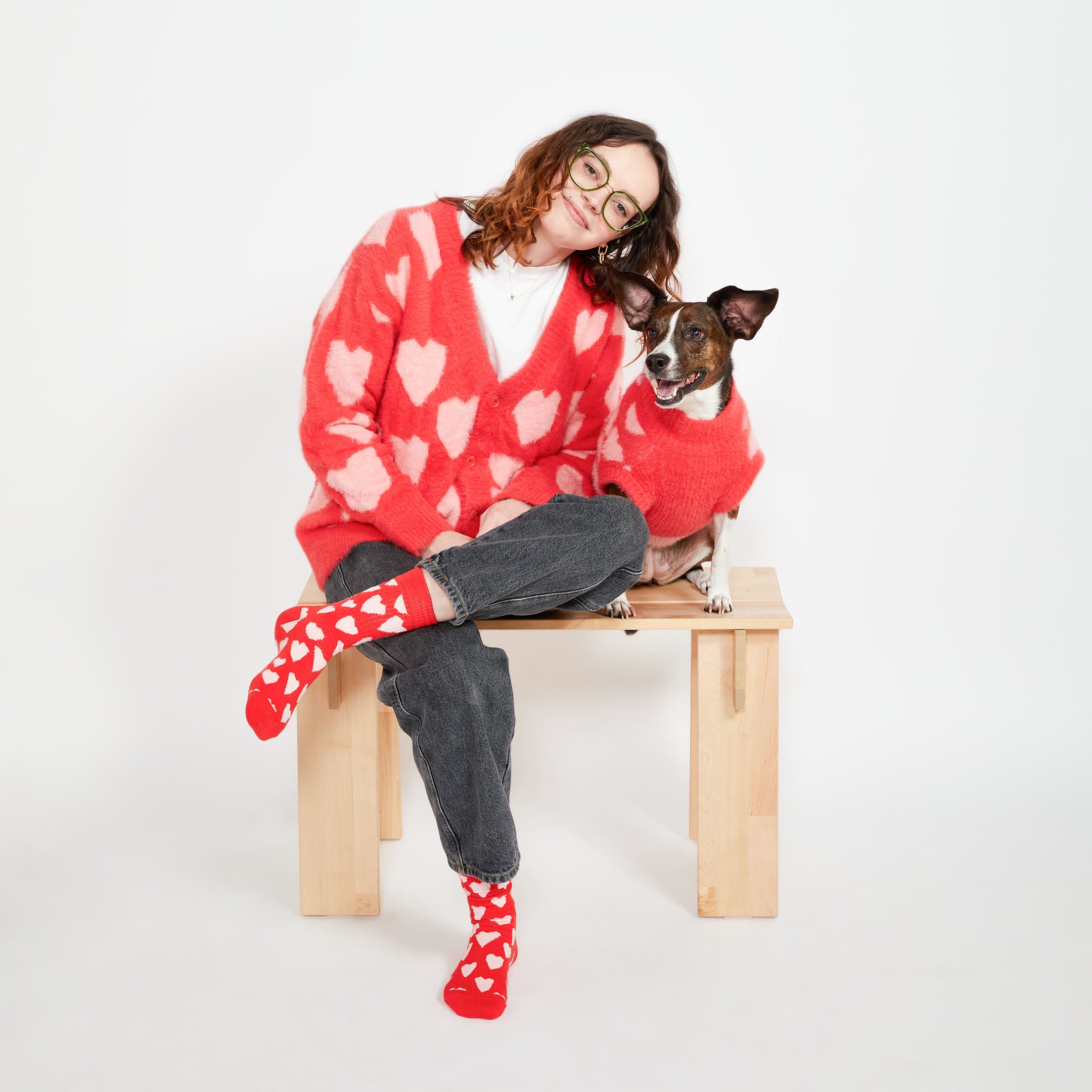 A woman sitting on a small wooden bench beside a brown dog. Both are wearing matching red sweaters with pink heart patterns, and the woman is wearing red socks with white hearts.