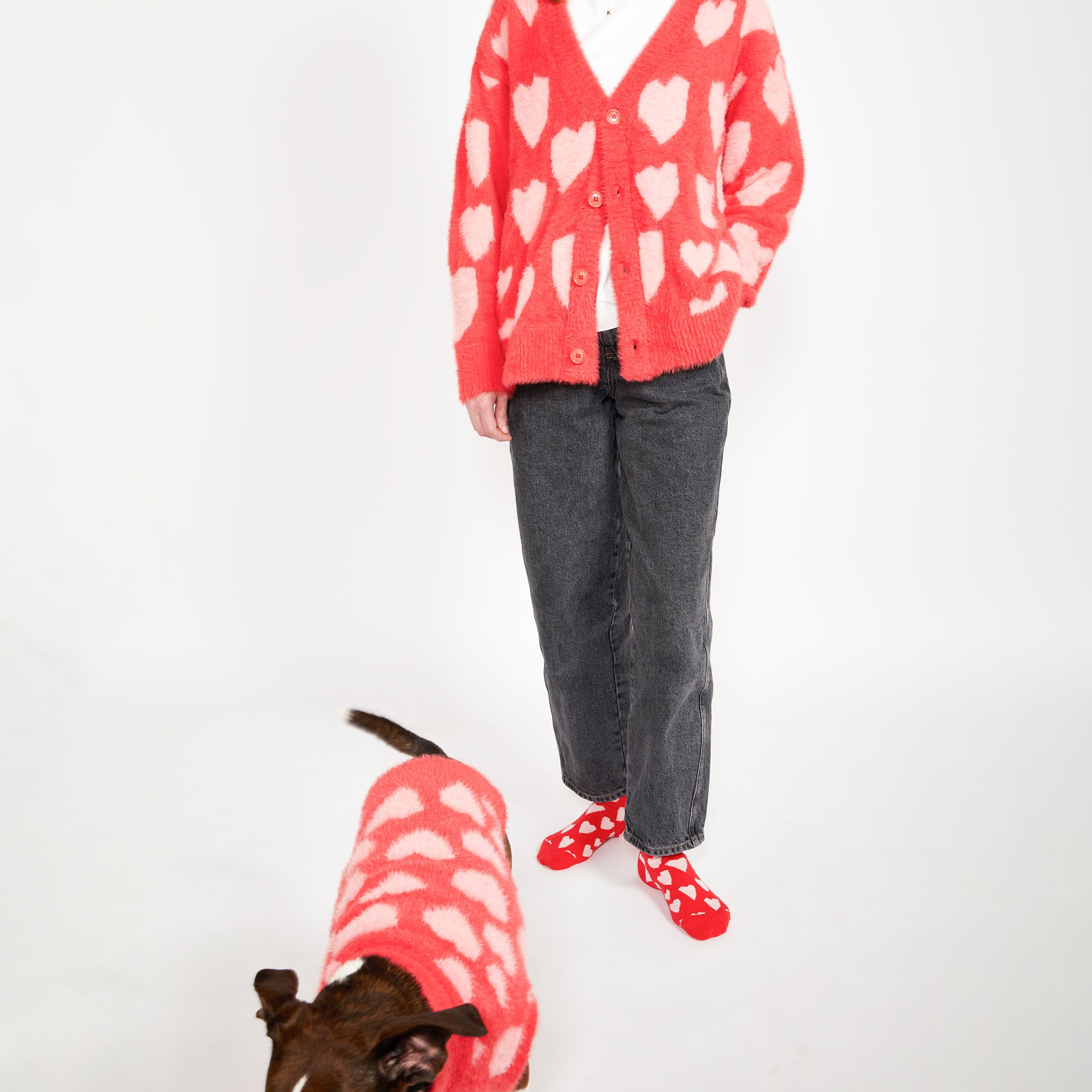 A woman standing in a red cardigan with pink hearts, paired with red socks with white hearts, while a brown dog wearing a matching red sweater walks nearby.