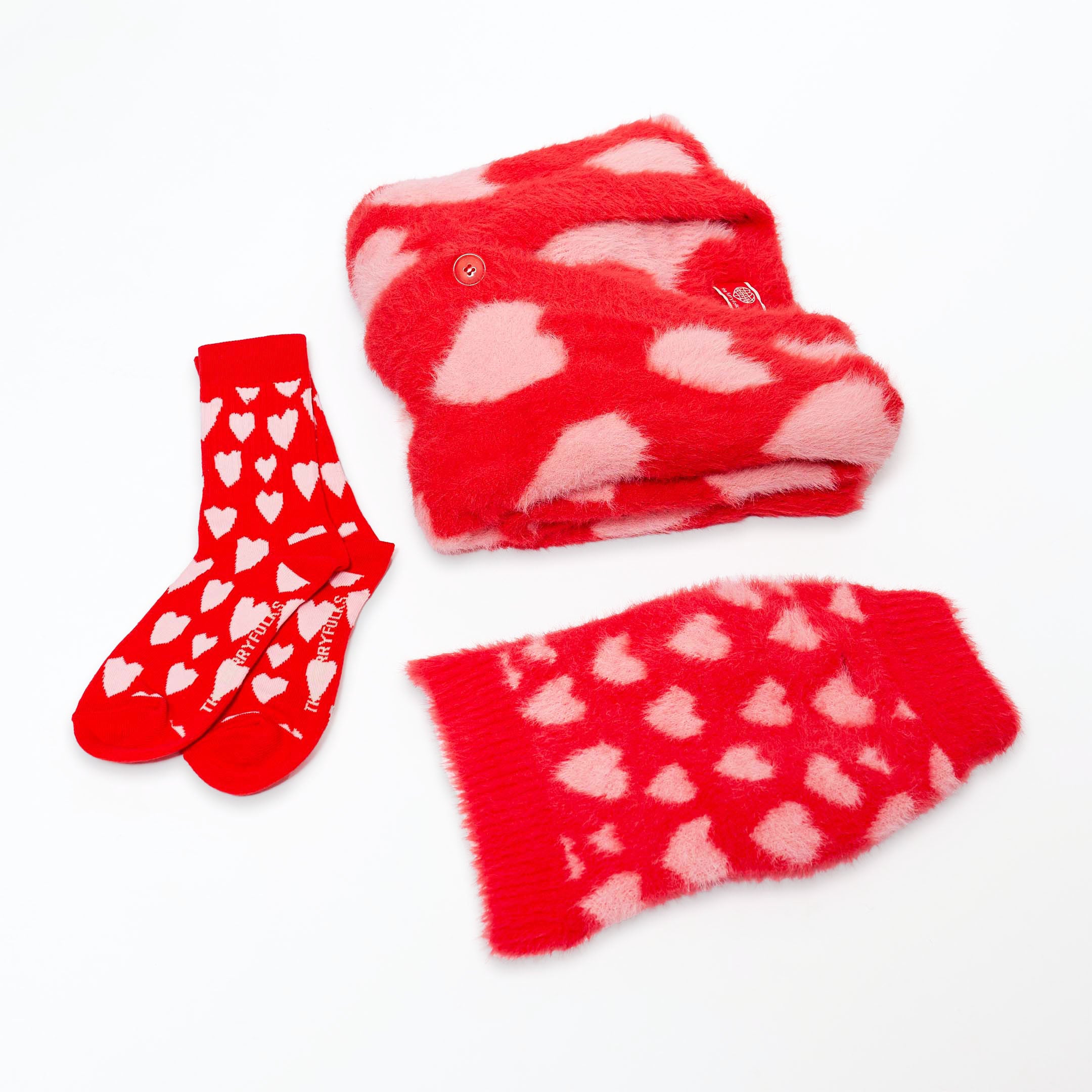 This image shows a red and pink heart-themed matching set. It includes a pair of red socks with white hearts and "THE FURRY FOLKS" written in red at the toe, a red fluffy sweater with pink heart patterns, and a matching heart-patterned sweater for pets. The design is vibrant and playful, with a soft, cozy texture, perfect for matching human and pet outfits.