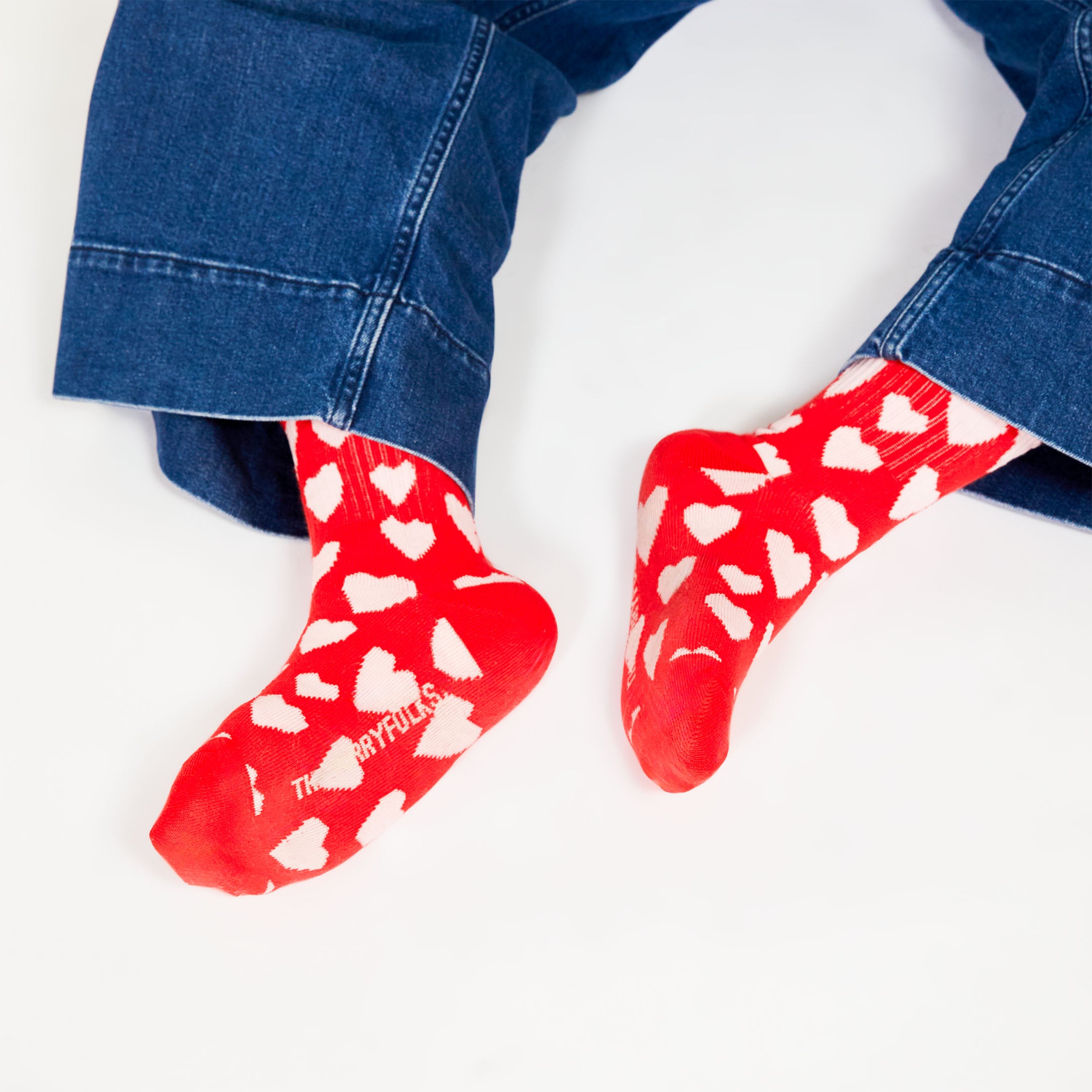  A close-up of red socks with white hearts, worn by someone seated, showing the playful heart pattern and "THEFURRYFOLKS" logo on the bottom of the socks.