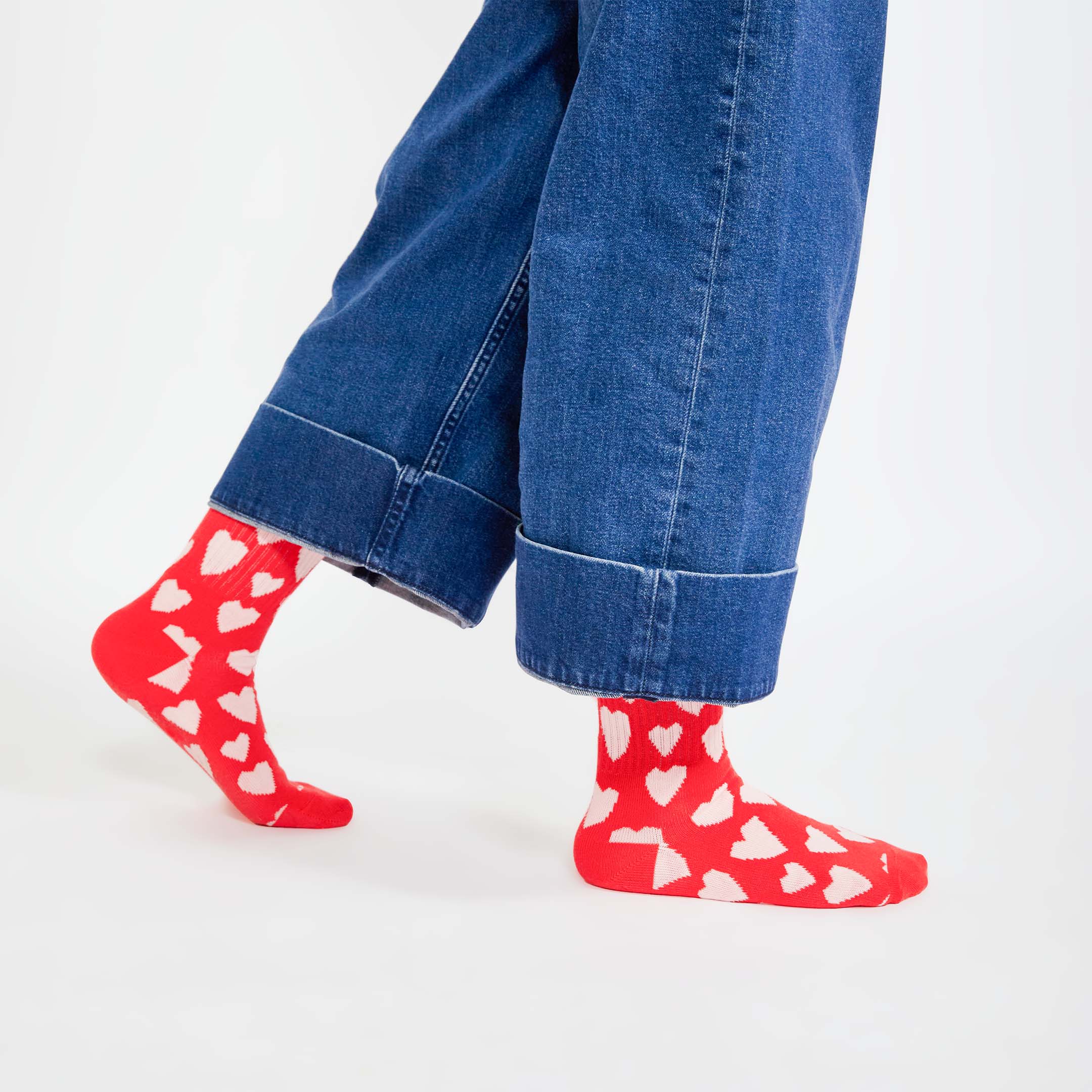 A close-up of someone walking, wearing wide-legged, cuffed denim jeans and red socks with white heart patterns. Only the lower legs and feet are visible, showcasing the playful heart design on the socks.