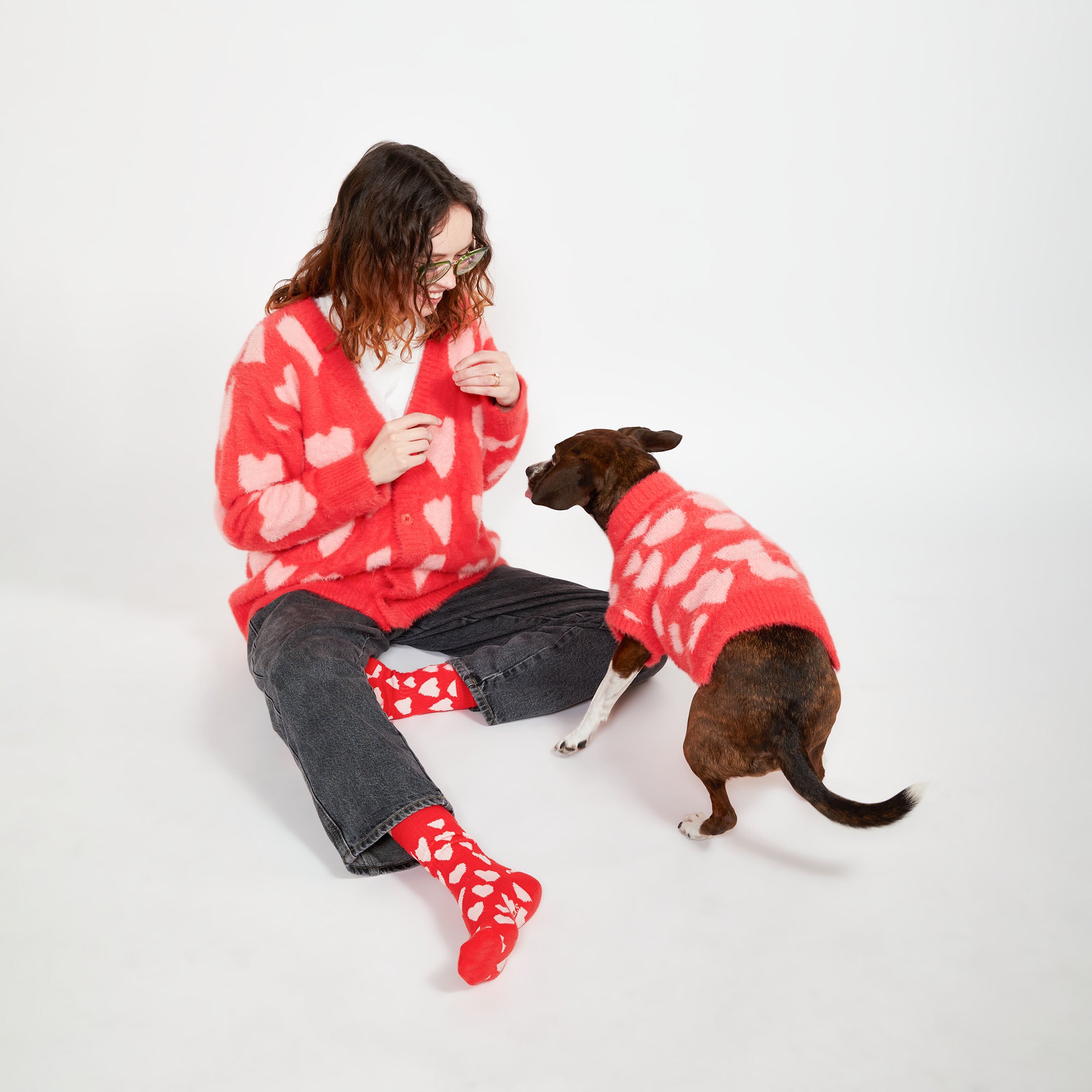  A woman sitting on the floor wearing a red cardigan with pink hearts, matching her dog who is wearing a red sweater with similar heart patterns. They both have matching heart-patterned socks.