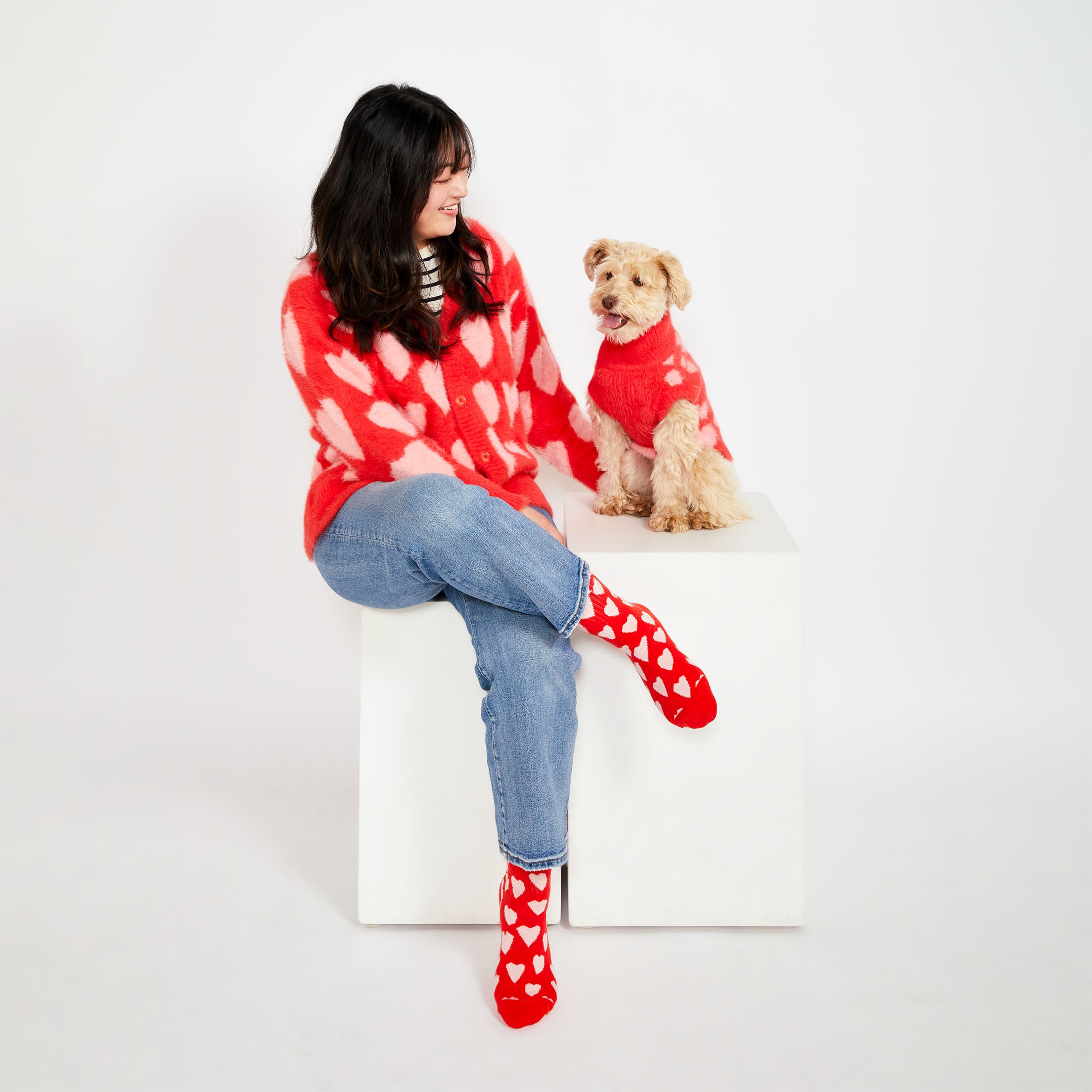 A woman seated on a white cube, wearing a red cardigan with pink hearts and matching red socks with white hearts. She is smiling at her small dog, who is also wearing a red sweater with pink heart patterns.