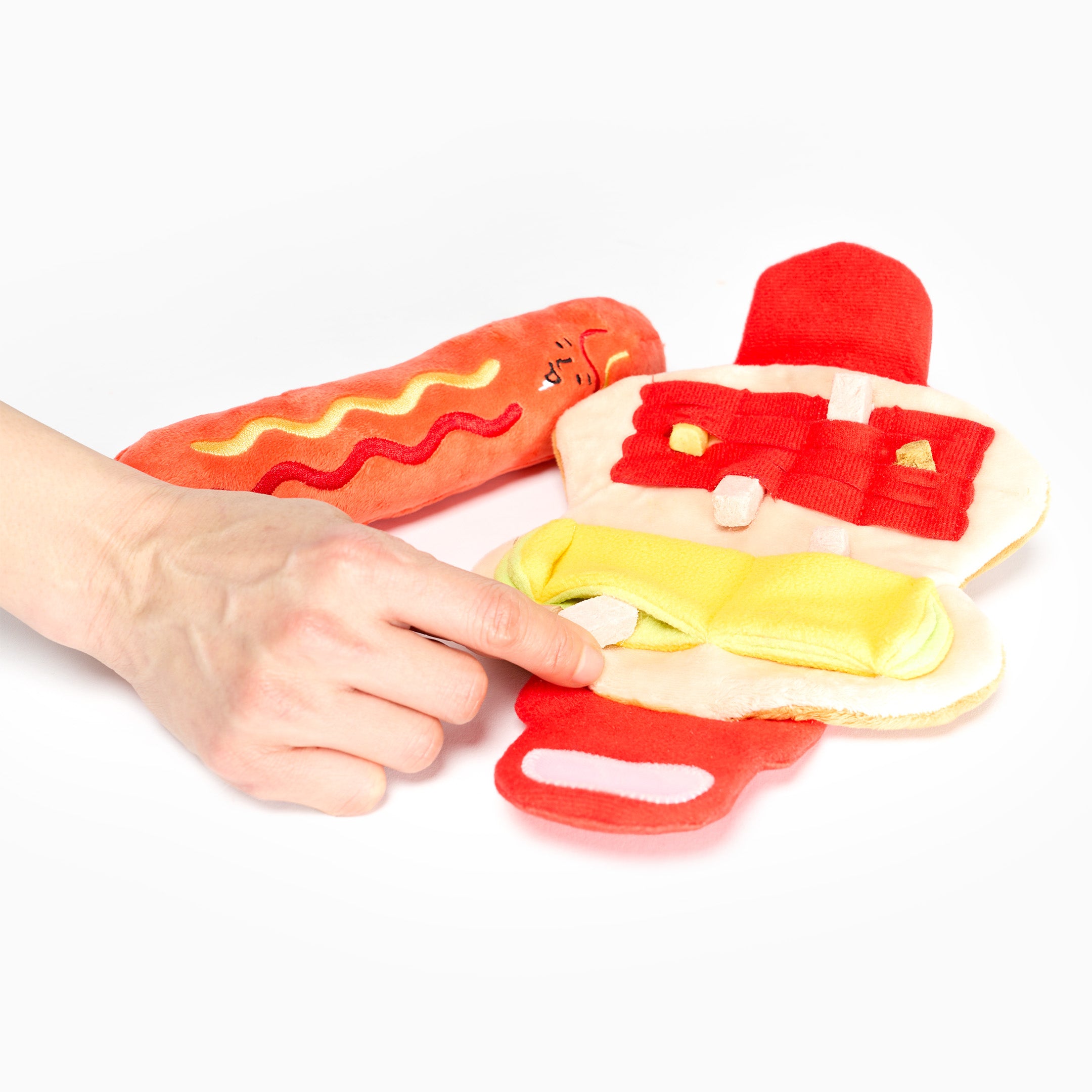 Hand holding a red and yellow toy hot dog with a bun on a white background