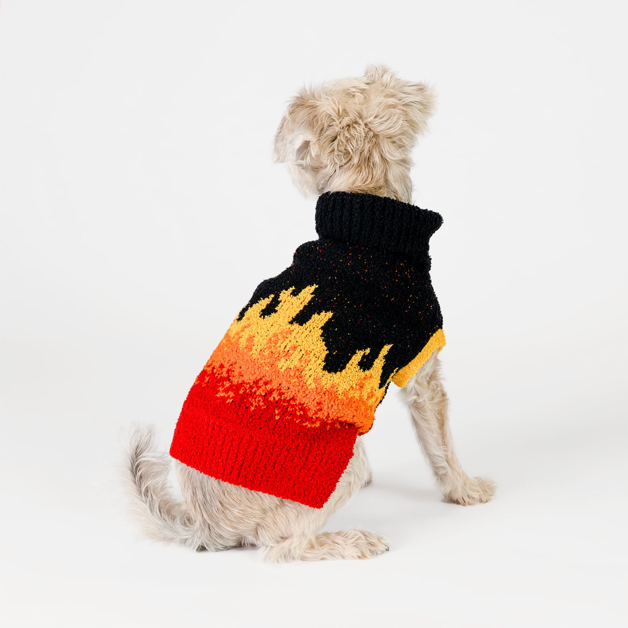 A small dog is sitting and facing away, showing off the back of its vibrant flame-patterned sweater. The sweater starts with a black turtleneck and gradually transitions into a striking flame design with yellow at the top, orange in the middle, and red at the bottom. The knit material looks soft and comfortable, covering the dog's body snugly while leaving its tail and legs free. 