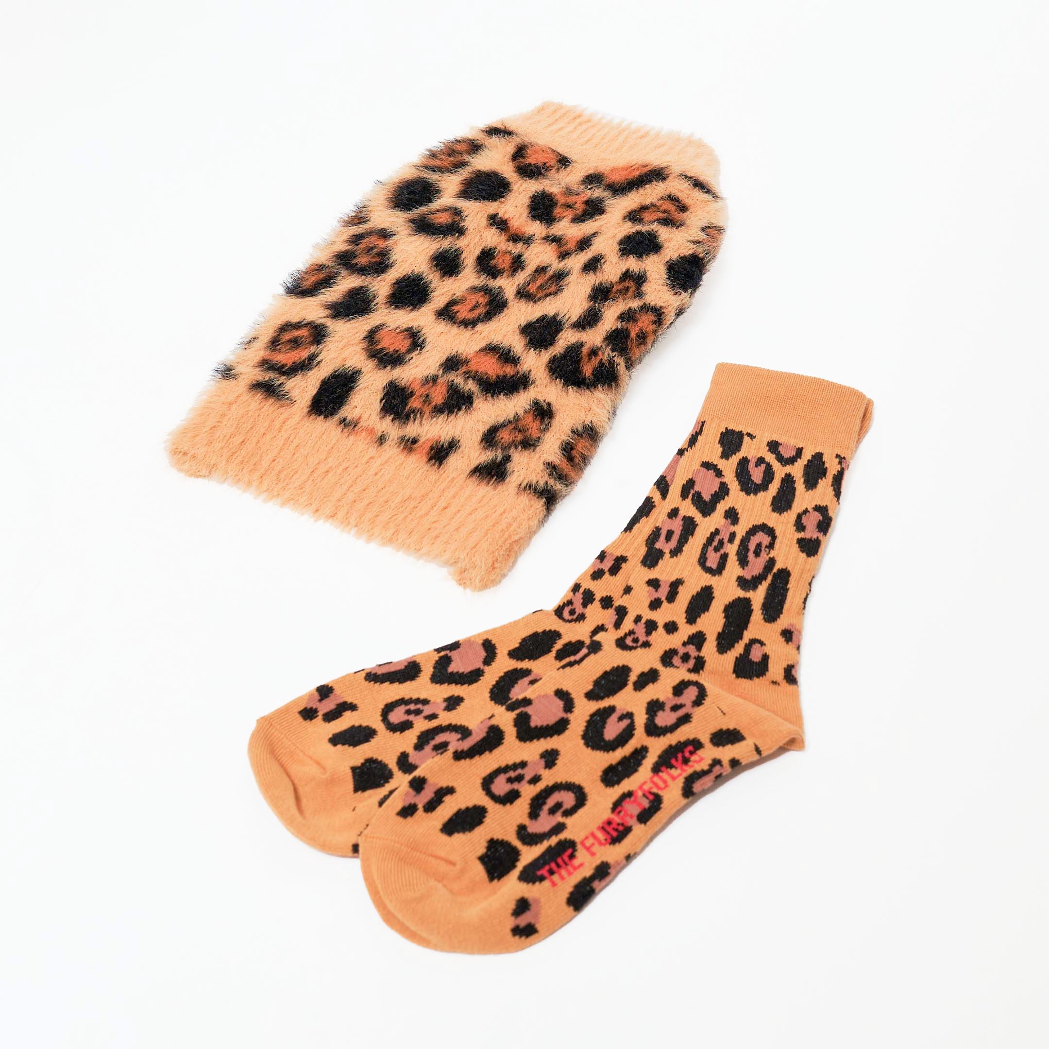 A pair of leopard-print socks is shown next to a matching leopard-print sweater. Both items feature a stylish, bold pattern with shades of brown, black, and tan. The socks have "THE FURRY FOLKS" written in red at the toe, indicating the brand. The sweater has a fluffy texture, giving it a cozy appearance. This is a matching set designed for both humans and pets.