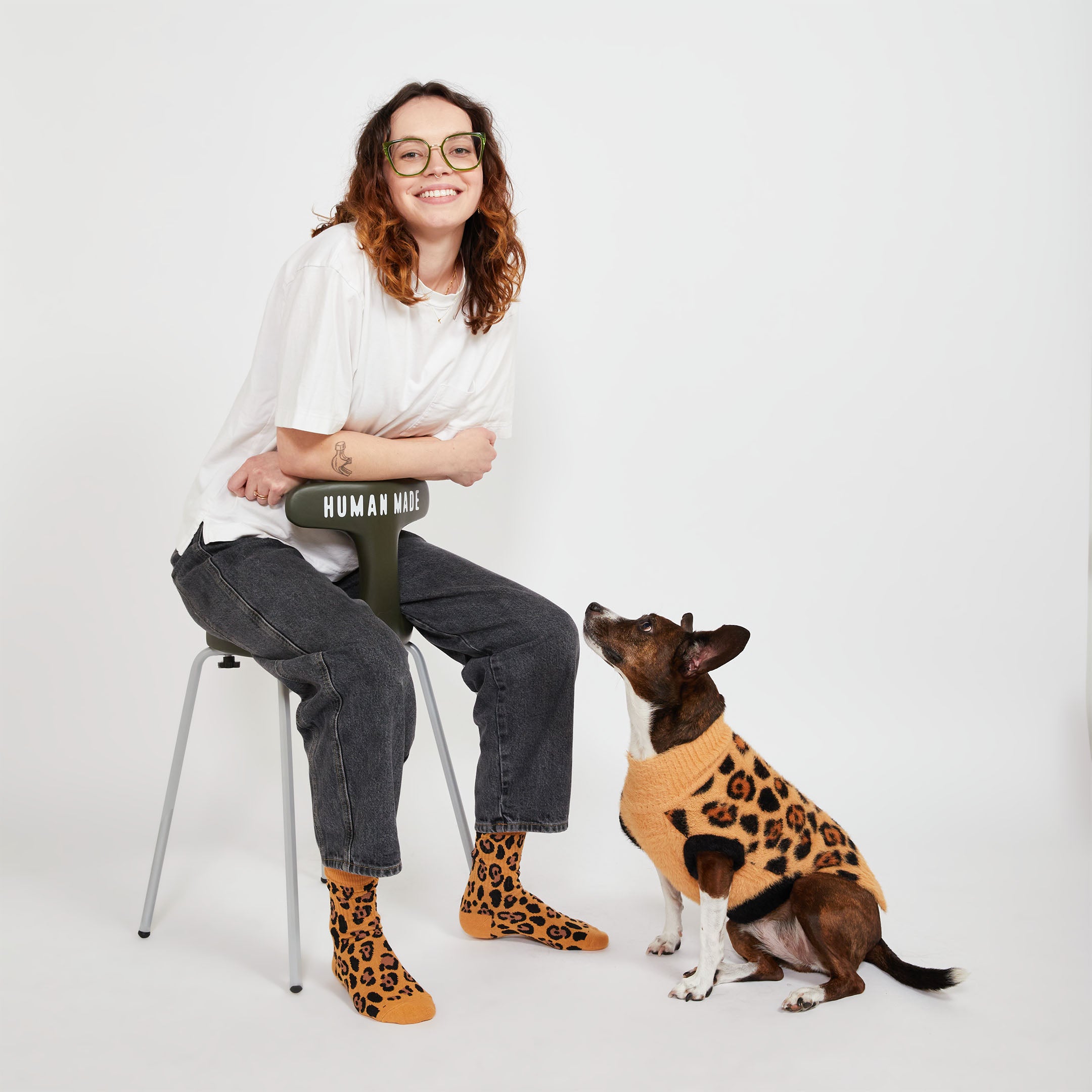 A person with glasses, sitting on a stool, smiles while looking at a dog in a leopard-print sweater. The dog is sitting and gazing back at the person. The person's socks match the dog's sweater in a leopard-print design.