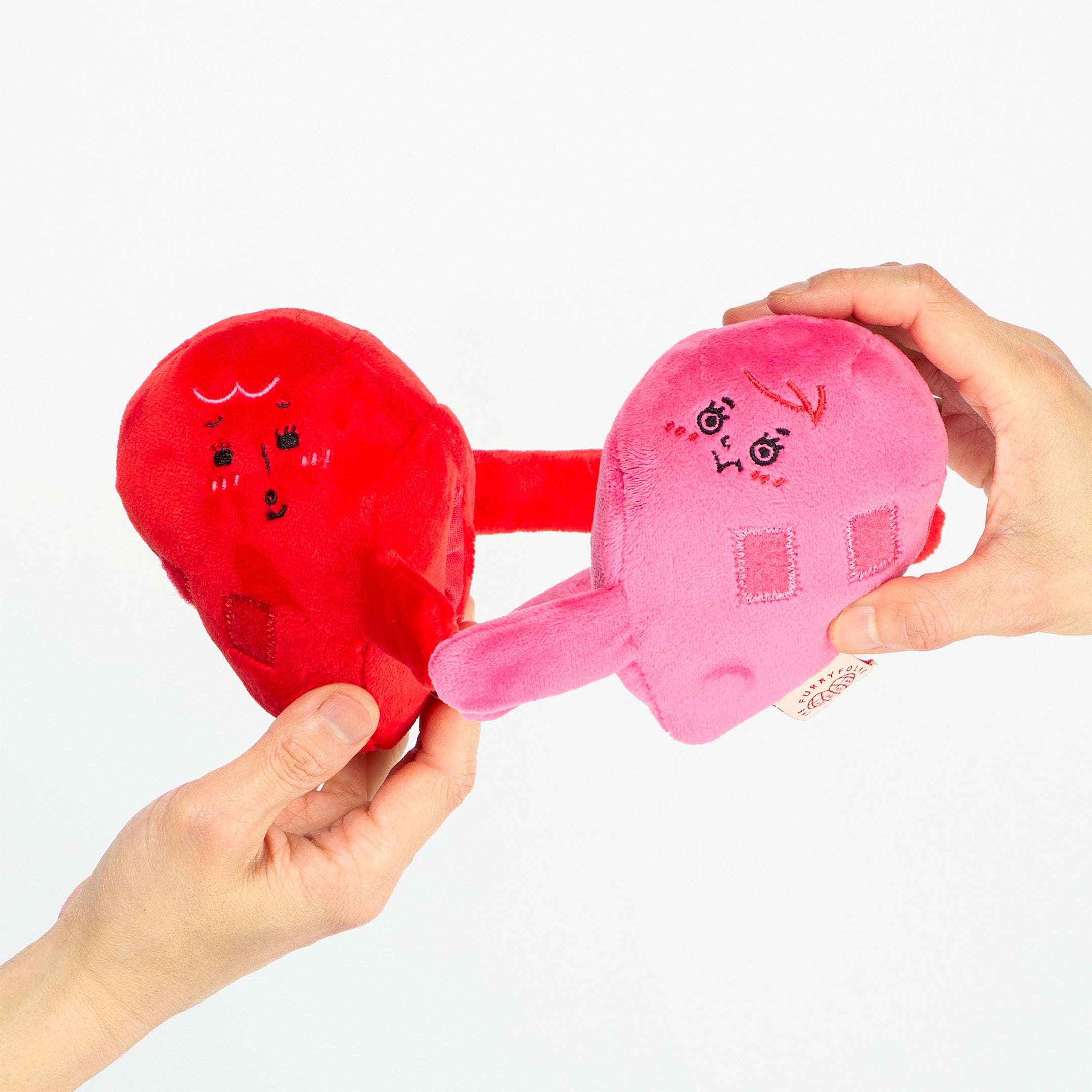 Two plush toys shaped like a red heart and pink house held by hands against a white background