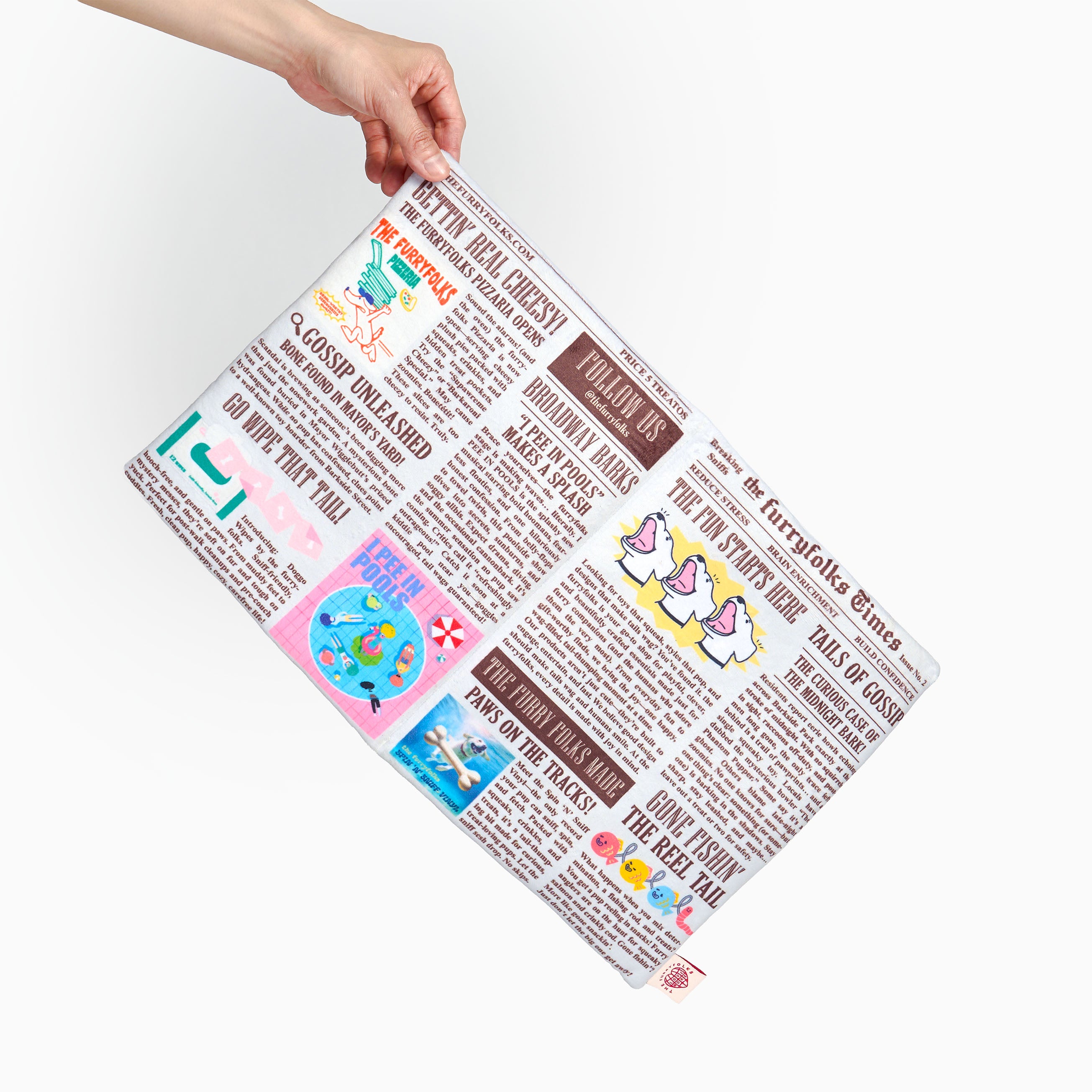 A hand holding a rectangular placemat printed with a colorful cartoon newspaper design, shown against a plain white background.