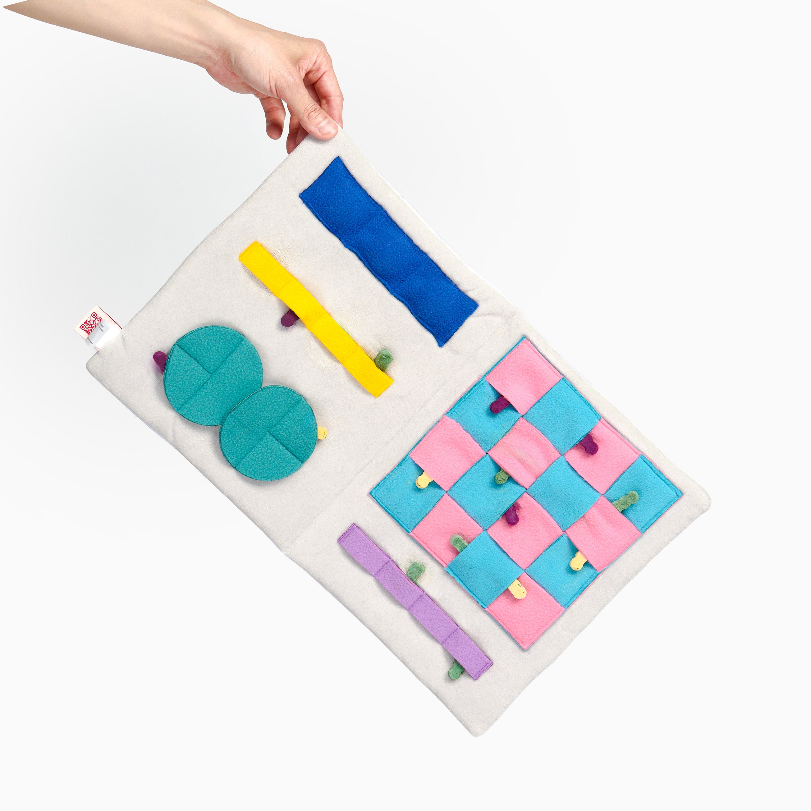 A fabric sensory activity mat held by a hand against a plain white background. The mat is off-white and features multiple colorful sensory tabs, including blue, yellow, teal, pink, and purple shapes.