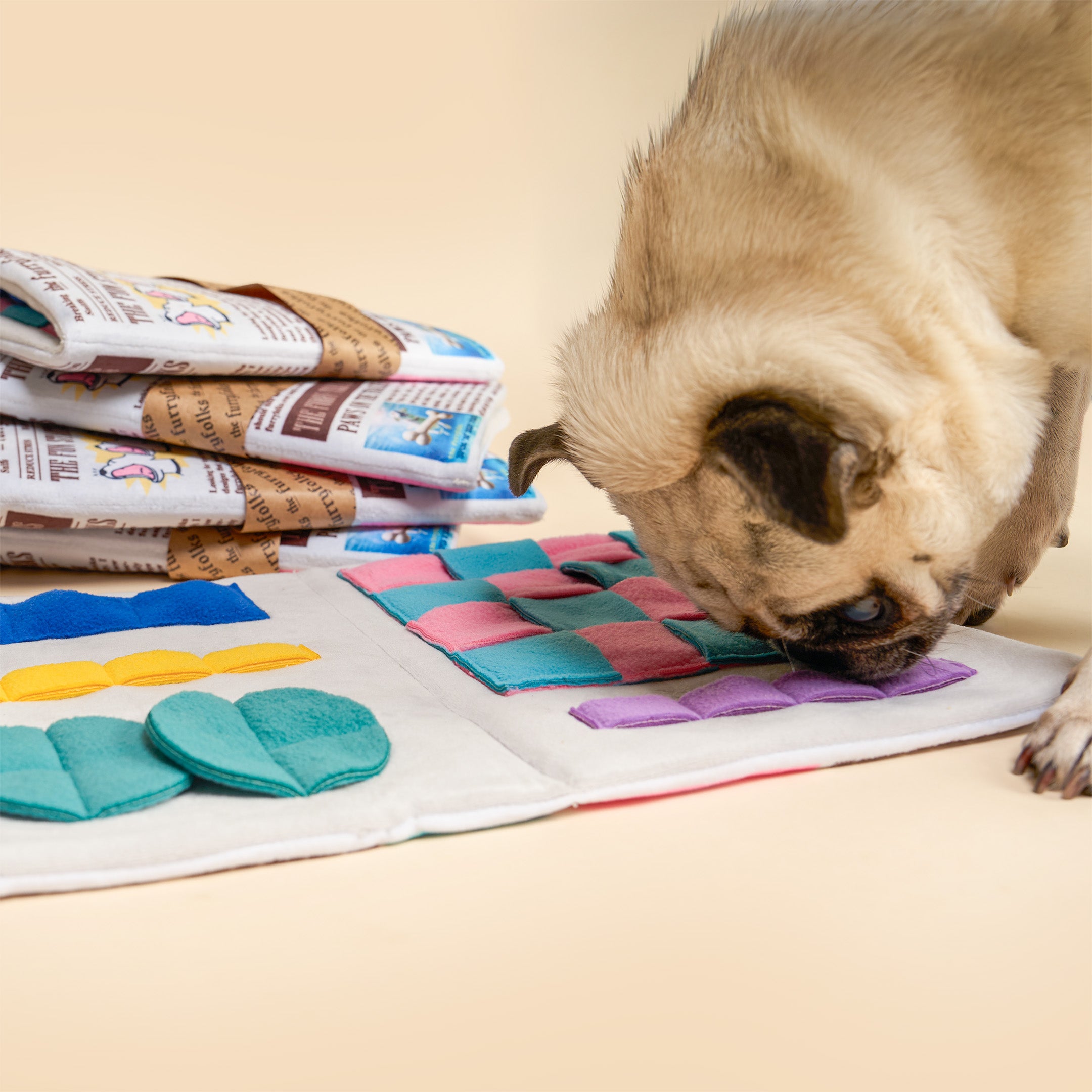A pug dog is sniffing a colorful felt puzzle mat on a beige surface, with a stack of folded puzzle mats in the background.