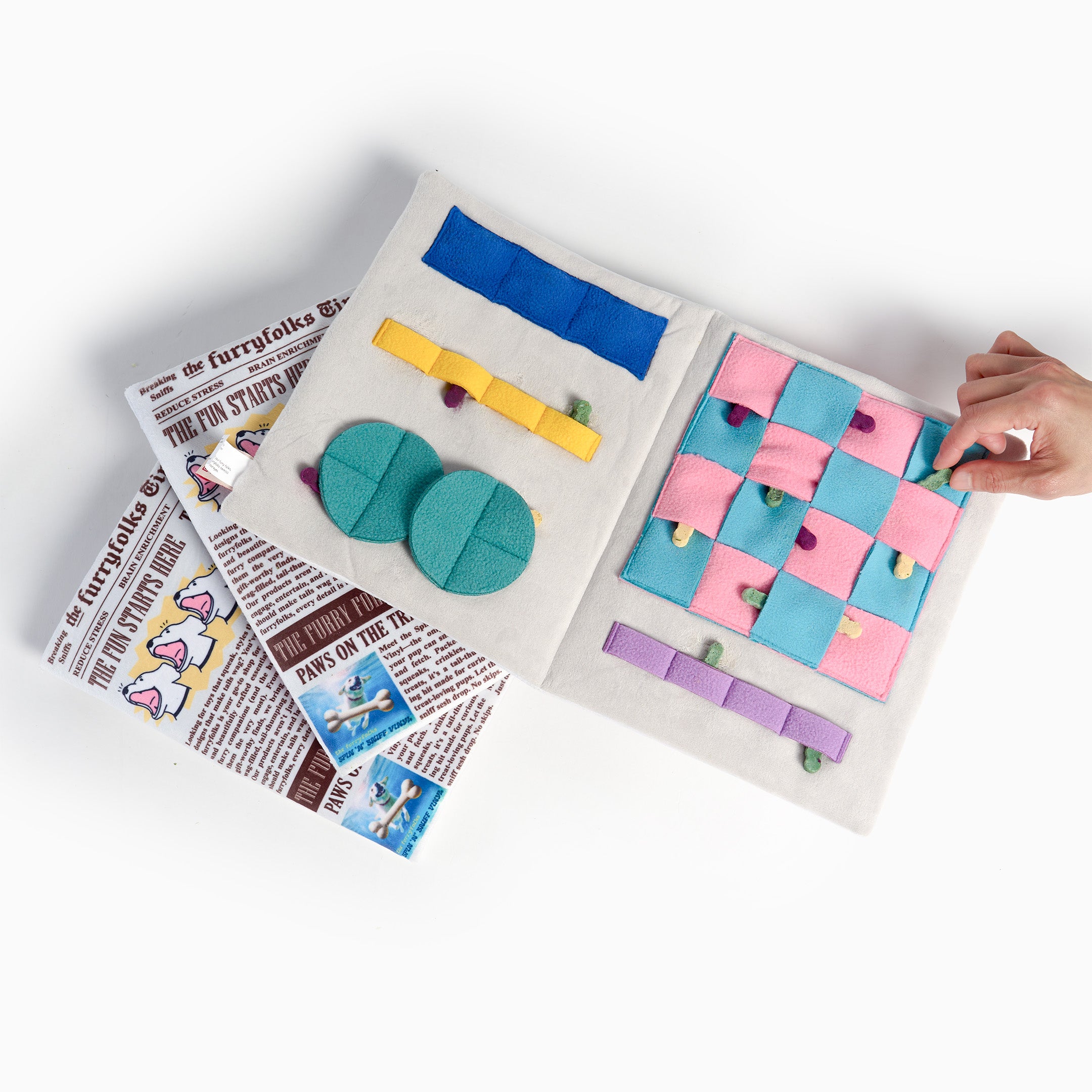 An open activity book with colorful felt tabs, buttons, and a checkered pattern panel, shown on a white background with a hand interacting with the pages. Newspaper pages are visible underneath as props.