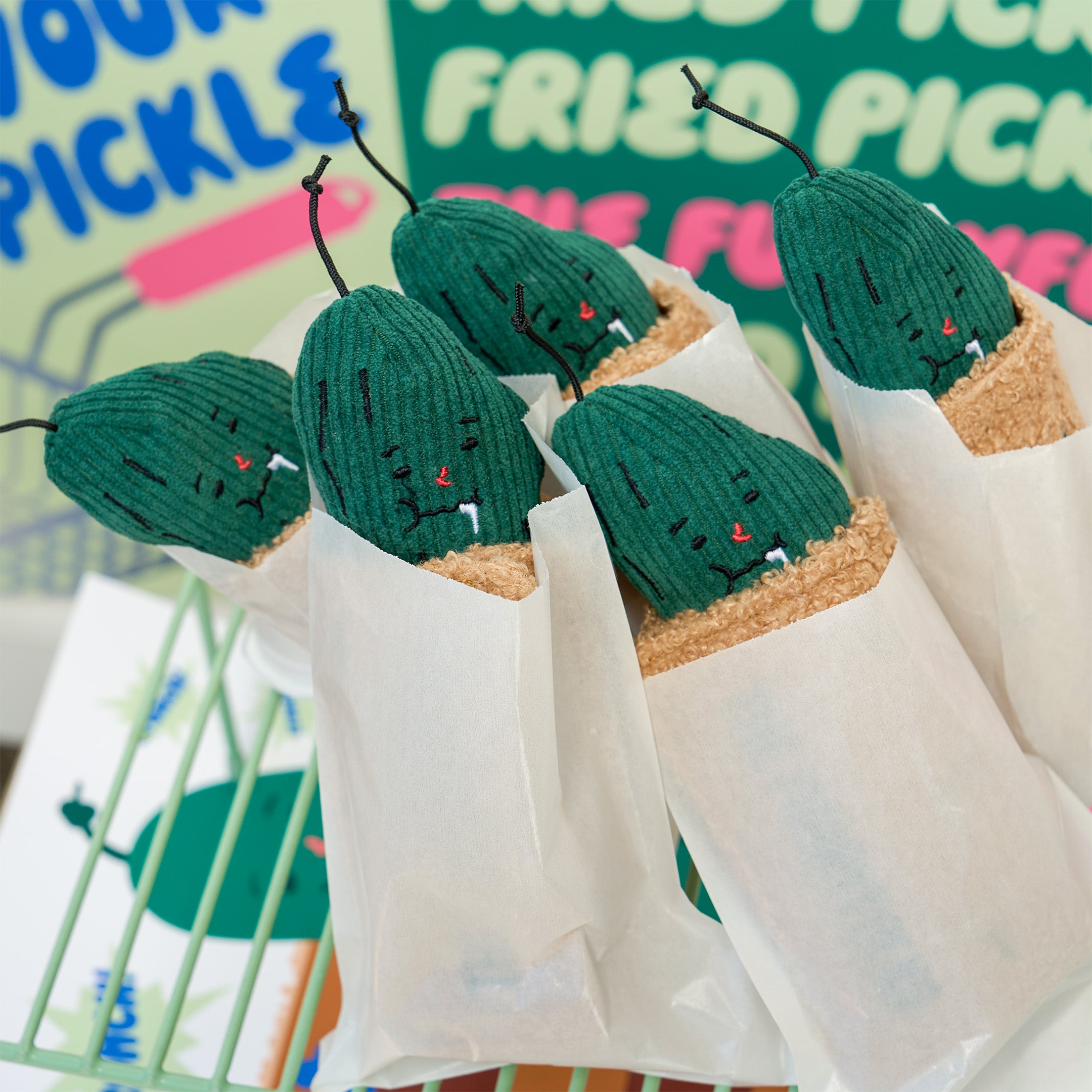 Five smiling, pickle-shaped plush dog toys sit upright in white paper bags against a colorful “fried pickles” display.