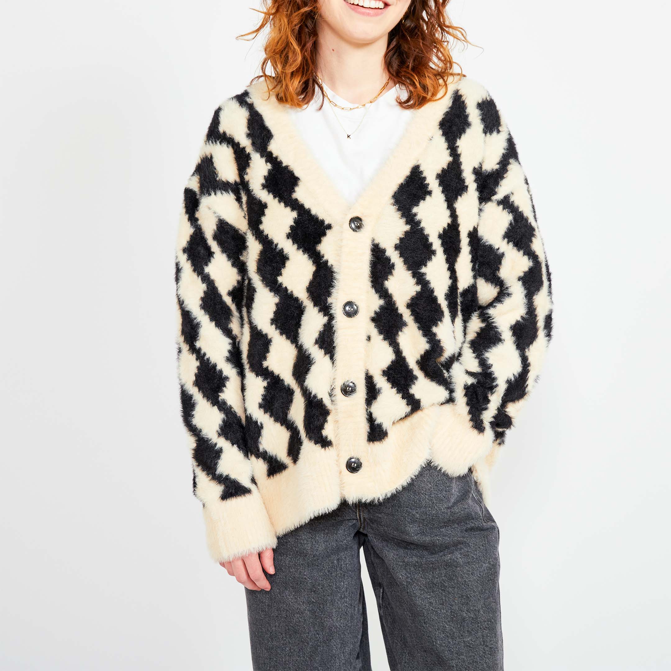 A close-up of a woman smiling while wearing a black and cream-colored cardigan with a bold diamond pattern. Her casual outfit includes dark jeans.