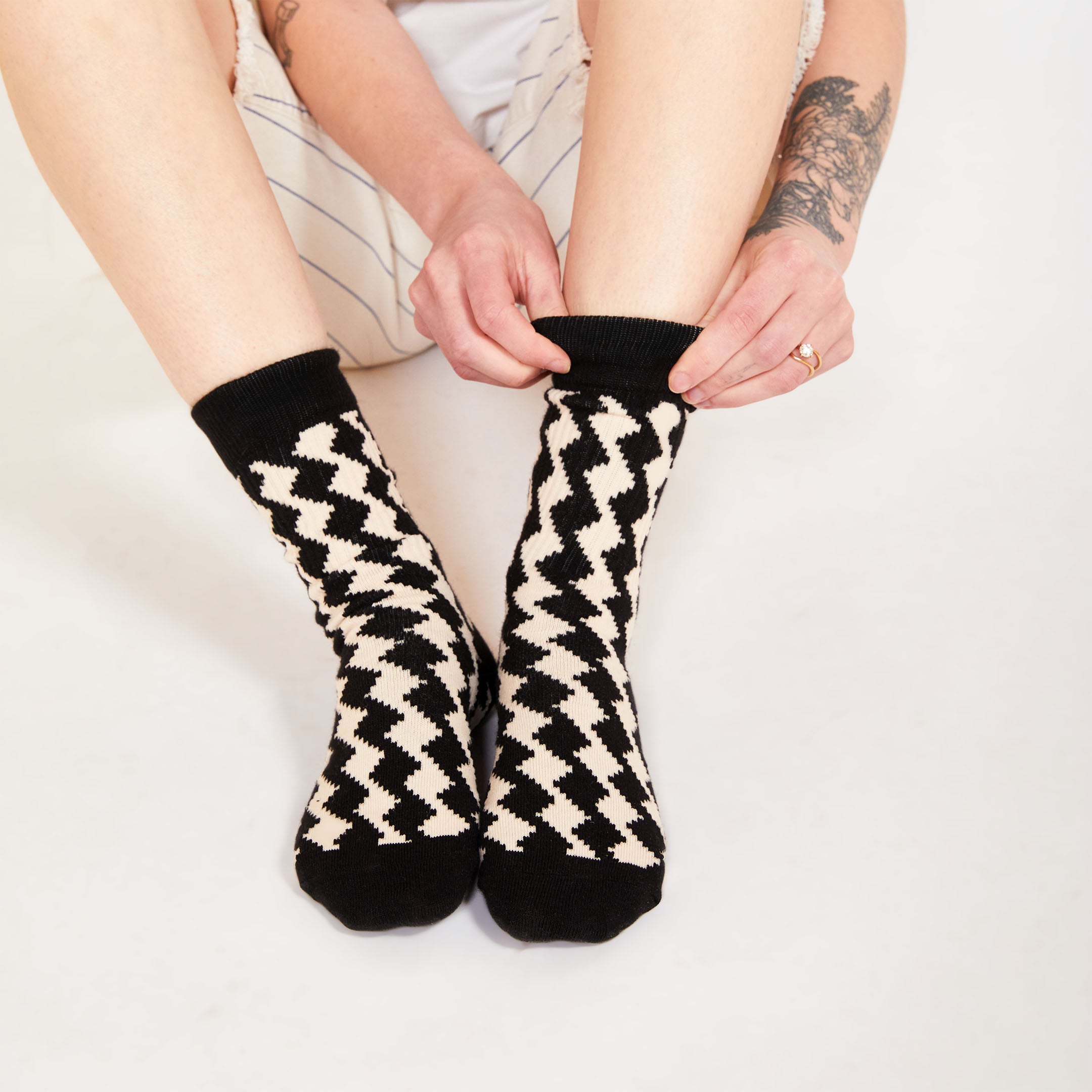 A close-up of a person adjusting their cream and black diamond-patterned socks, sitting on the floor with hands pulling the top of the socks up.