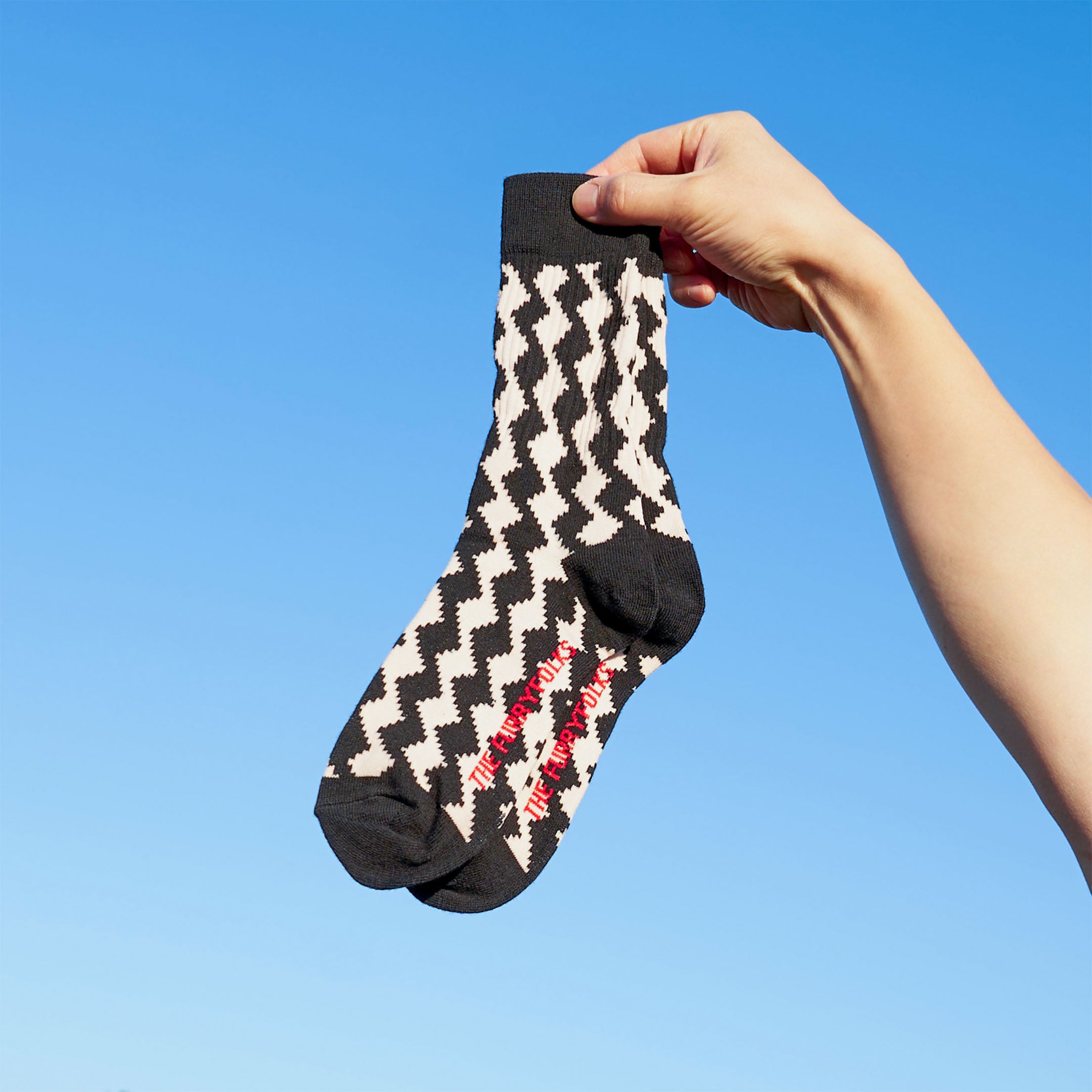 A hand holding a pair of black and cream diamond-patterned socks against a bright blue sky. The socks have "THE FURRYFOLKS" logo written on the bottom in red.