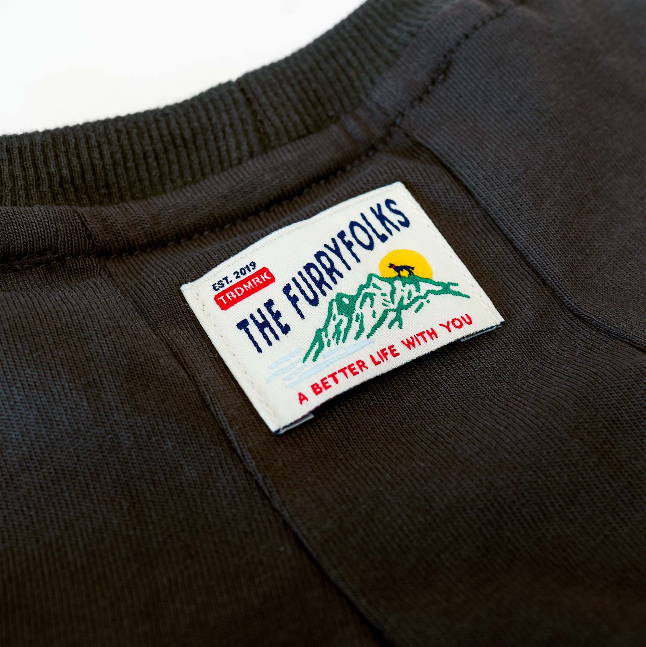 Close-up image of a clothing label on a dark fabric, featuring the logo and tagline of the furryfolks. The label shows an illustrated mountain landscape with a sun and an animal silhouette, accompanied by the text ‘EST. 2019, TRADEMARK, THE FURRYFOLKS’ and the tagline ‘A BETTER LIFE WITH YOU.