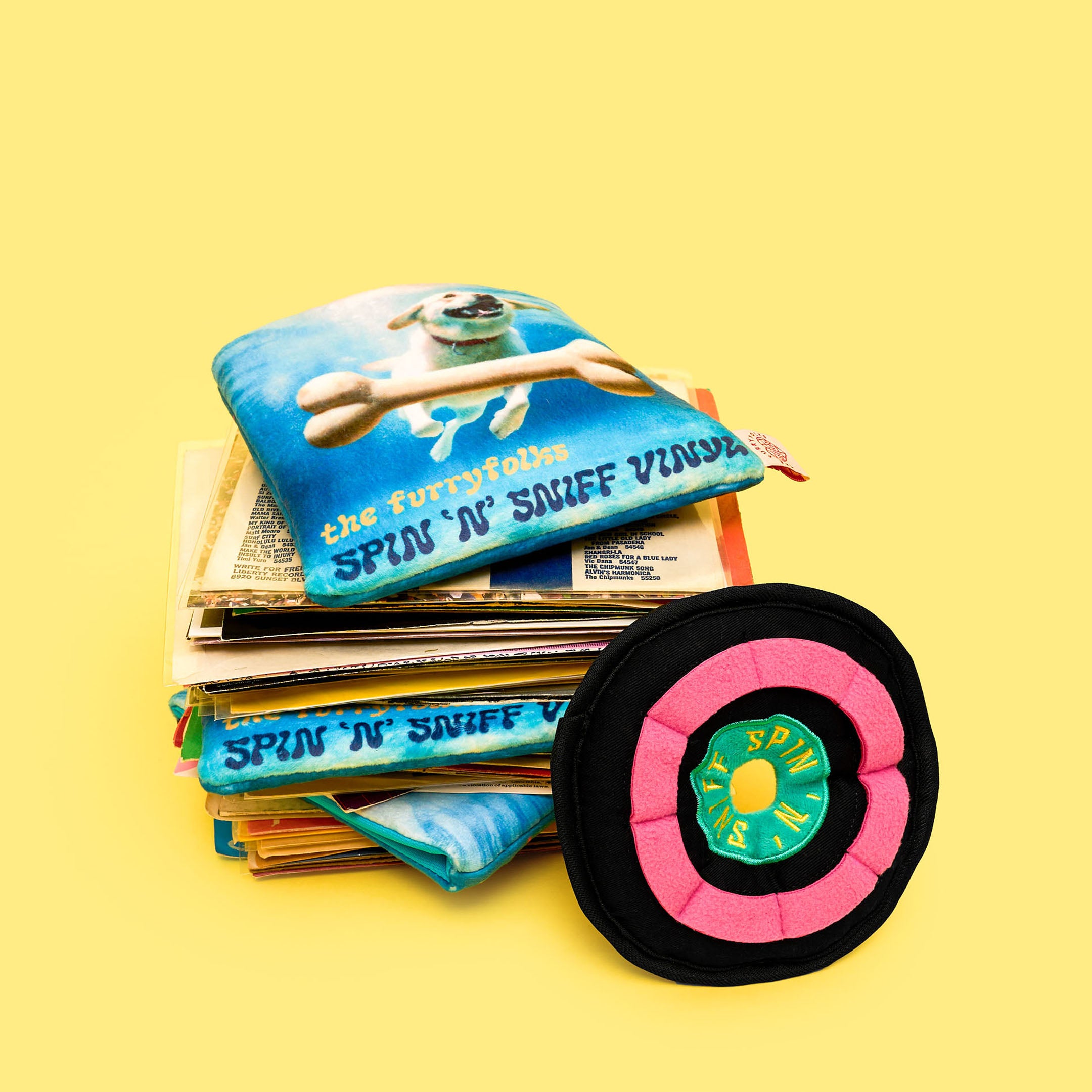 A stack of vinyl records topped with the furryfolks’ crinkly record sleeve and plush vinyl nosework toy in pink and black labeled “Spin ‘N’ Sniff.”