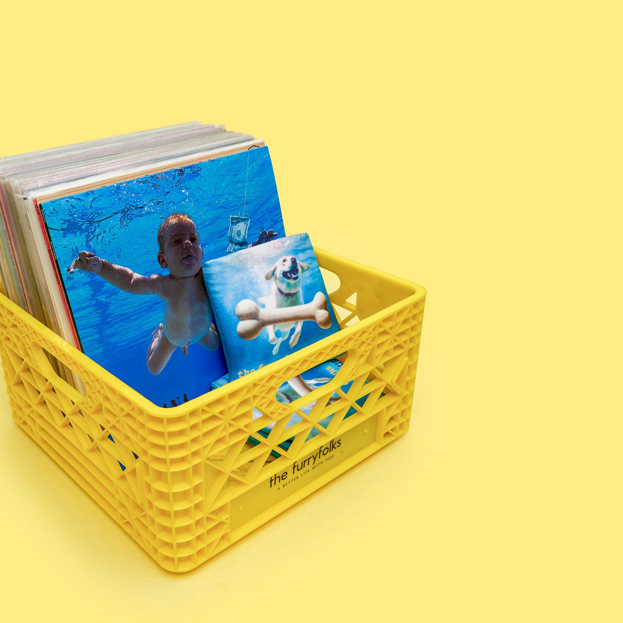 A yellow milk crate filled with vinyl records, including a parody dog toy album cover from the furryfolks featuring a dog underwater with a bone, mimicking Nirvana’s “Nevermind.”