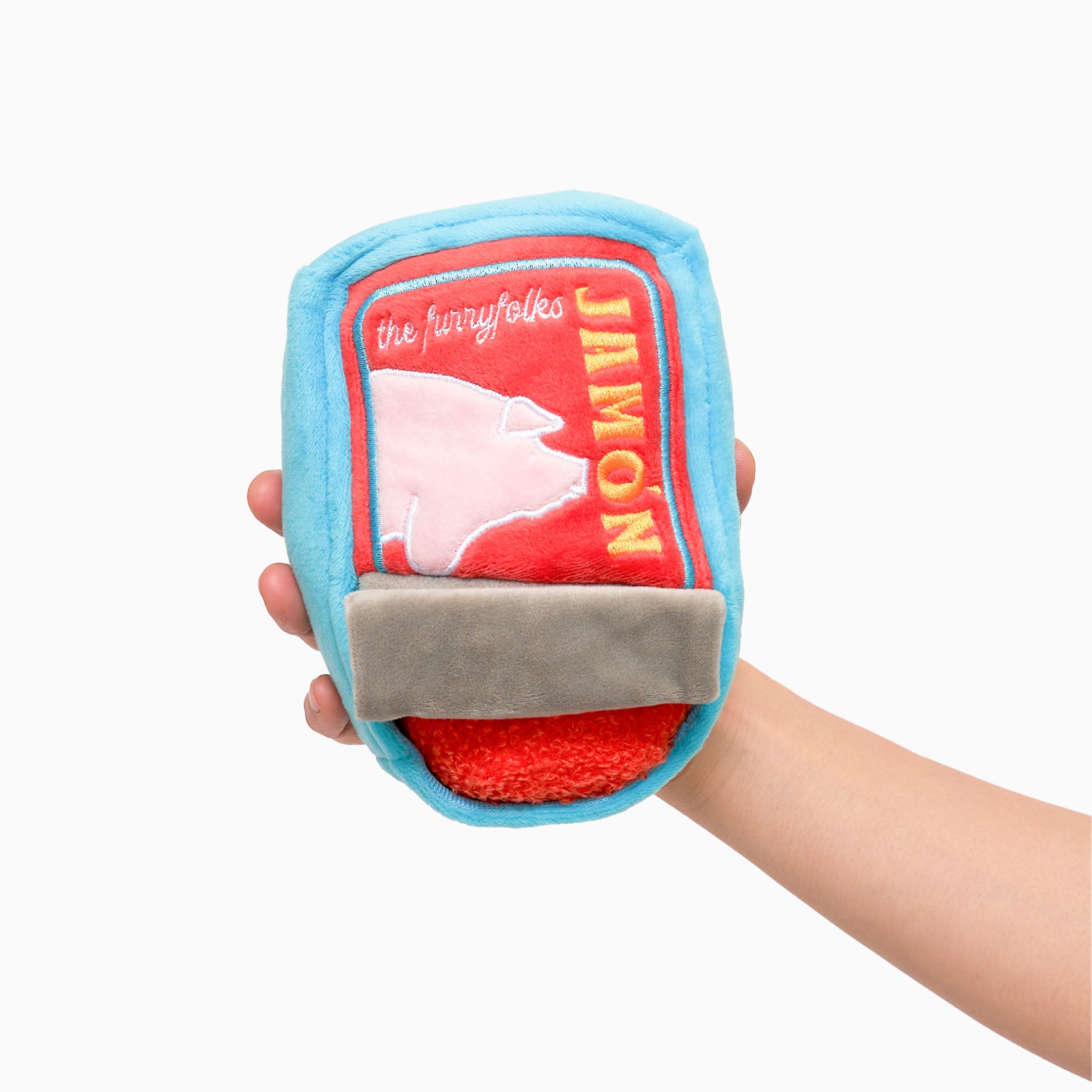 The image shows a hand holding a plush dog toy designed to look like a tin of jamón (ham). The toy has a bright blue exterior with a red label on the front. The label features a silhouette of a pig and the word “JAMÓN” in yellow letters. The bottom part of the toy has a gray section that looks like a pull tab. The toy is soft and designed for nosework activities for dogs, providing both entertainment and mental stimulation.