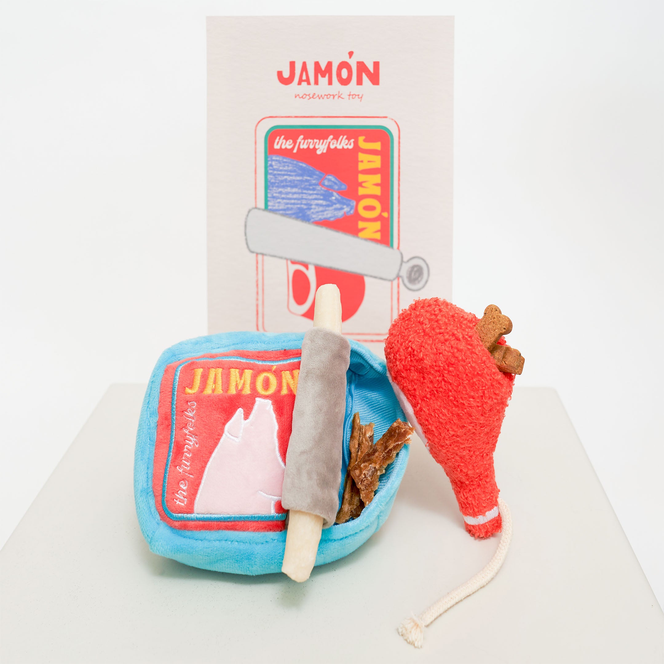 The image shows a dog nosework toy set called “Jamón.” The set includes a blue plush container designed to look like a tin can with a red label featuring a pig’s head and the words “Jamón” and “the furryfolks.” Inside the container are various plush components, including a red drumstick-shaped toy with brown bones sticking out and a grey roller toy. The background has a card with similar “Jamón” branding and a colorful illustration of the tin can with a roller and bones.