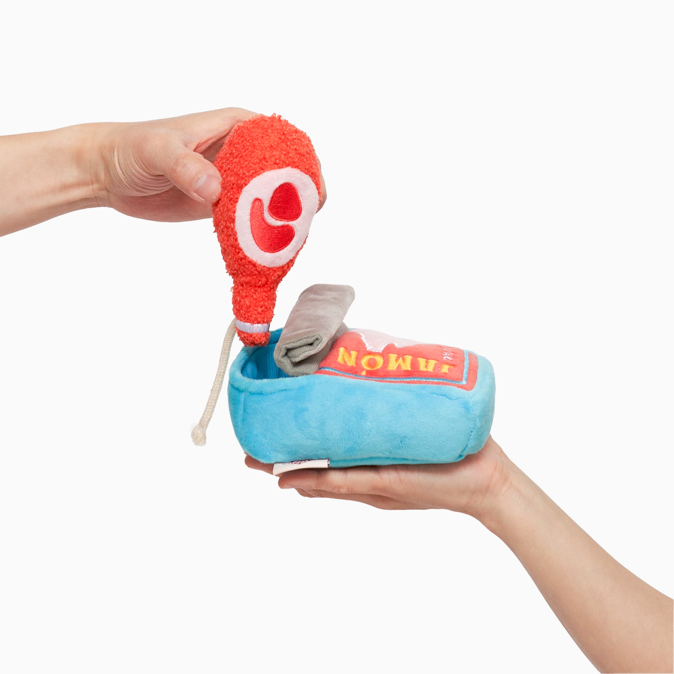 A hand is holding a plush dog toy shaped like a can of jamón (ham). The toy has a blue body with a red front label featuring a pink pig silhouette and the word “JAMÓN” in yellow. There’s a gray flap at the bottom resembling a can tab. The toy is soft and designed for dogs’ nosework activities.