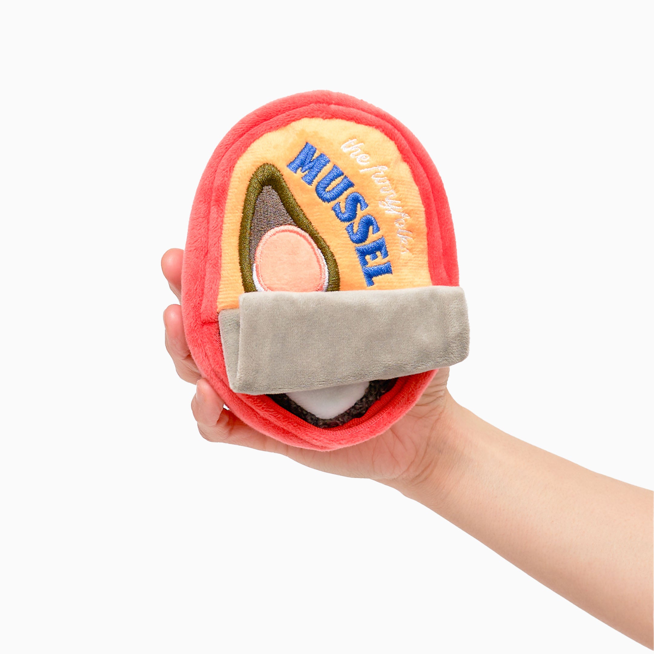 A hand is holding a plush dog toy shaped like a can of mussels. The toy has a red exterior with a yellow label on the front. The label features an illustration of a mussel and the word “MUSSEL” in blue letters. There is a gray flap at the bottom that resembles a pull tab. The toy is soft and designed for nosework activities for dogs, providing both entertainment and mental stimulation.