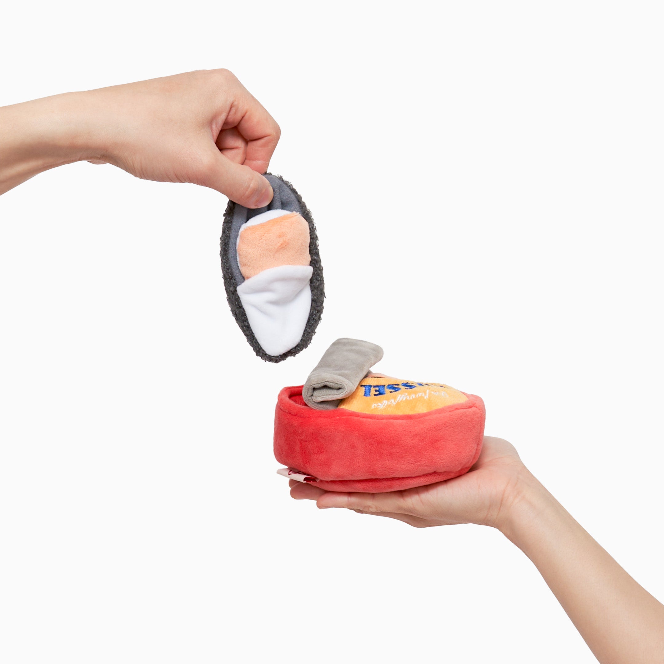 One hand is holding a plush toy shaped like a can of mussels, which is red with a yellow label. The label features an illustration of a mussel and the word “MUSSEL” in blue letters. Another hand is lifting a plush mussel from the can toy. The mussel plush is gray with a white and orange interior, mimicking the look of a real mussel. The toy is designed for dog nosework activities, providing mental stimulation and entertainment for pets.