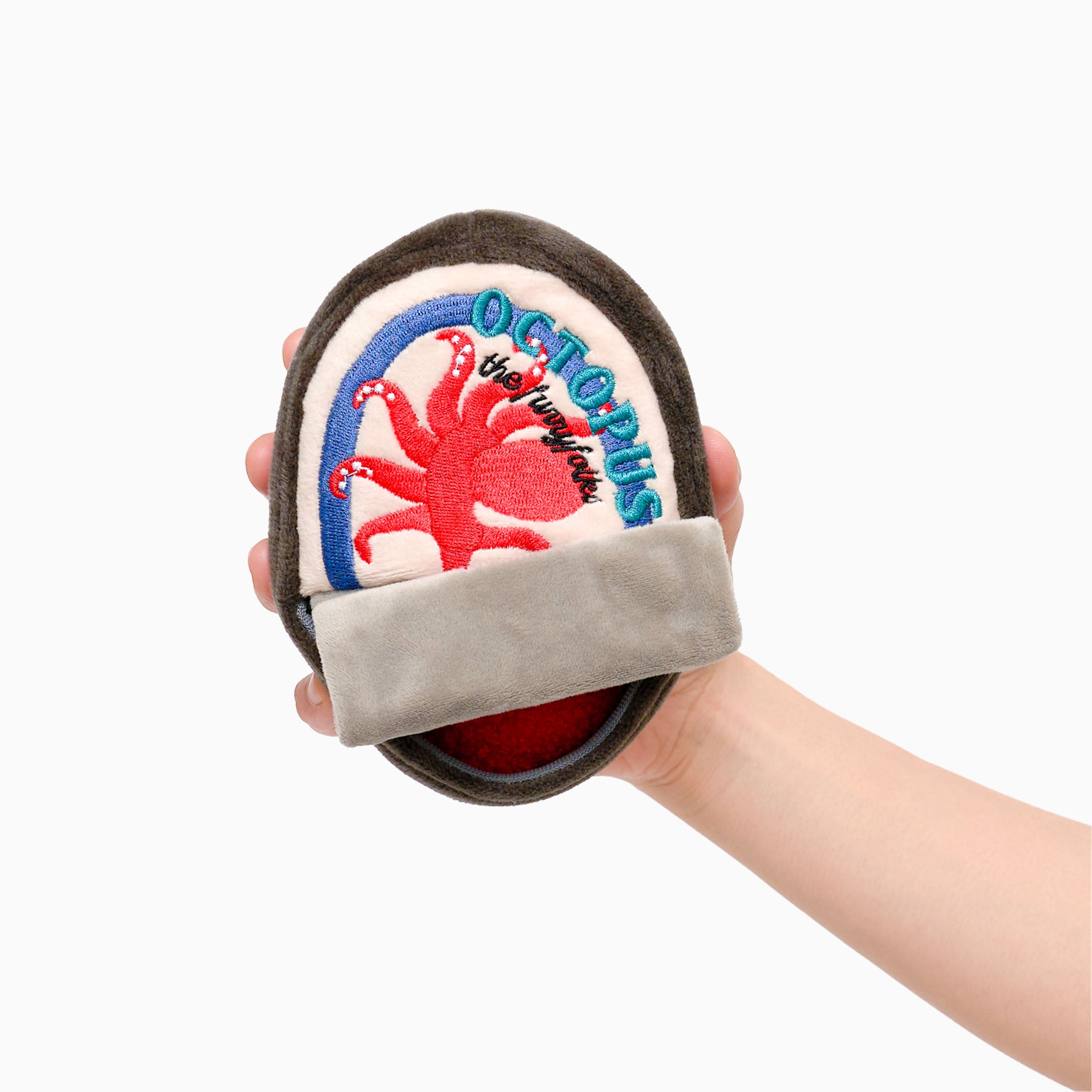 A hand is holding a plush dog toy shaped like a can of octopus. The toy has a dark brown exterior with a cream-colored label on the front. The label features a red octopus illustration and the word “OCTOPUS” in blue letters. There is a gray flap at the bottom that resembles a pull tab. The toy is soft and designed for nosework activities for dogs, providing both entertainment and mental stimulation.