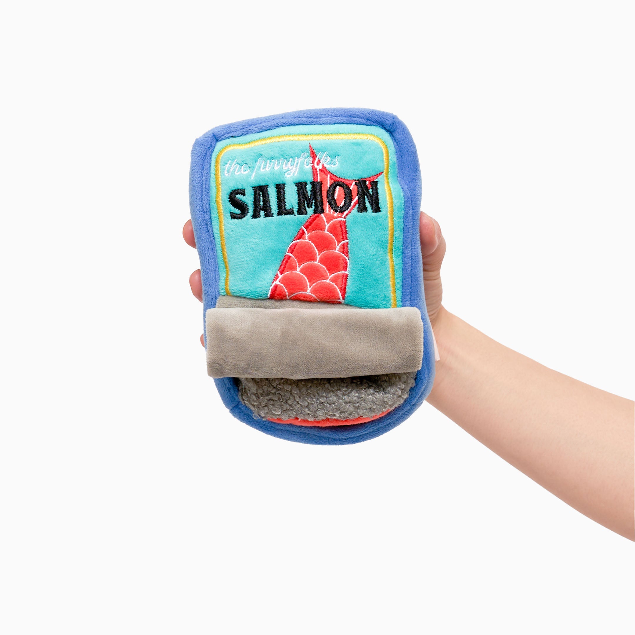 A hand is holding a plush dog toy shaped like a can of salmon. The toy has a blue exterior with a light blue label on the front. The label features an illustration of a red fish tail with scales and the word “SALMON” in black letters. There is a gray flap at the bottom that resembles a pull tab. The toy is soft and designed for nosework activities for dogs, providing both entertainment and mental stimulation.