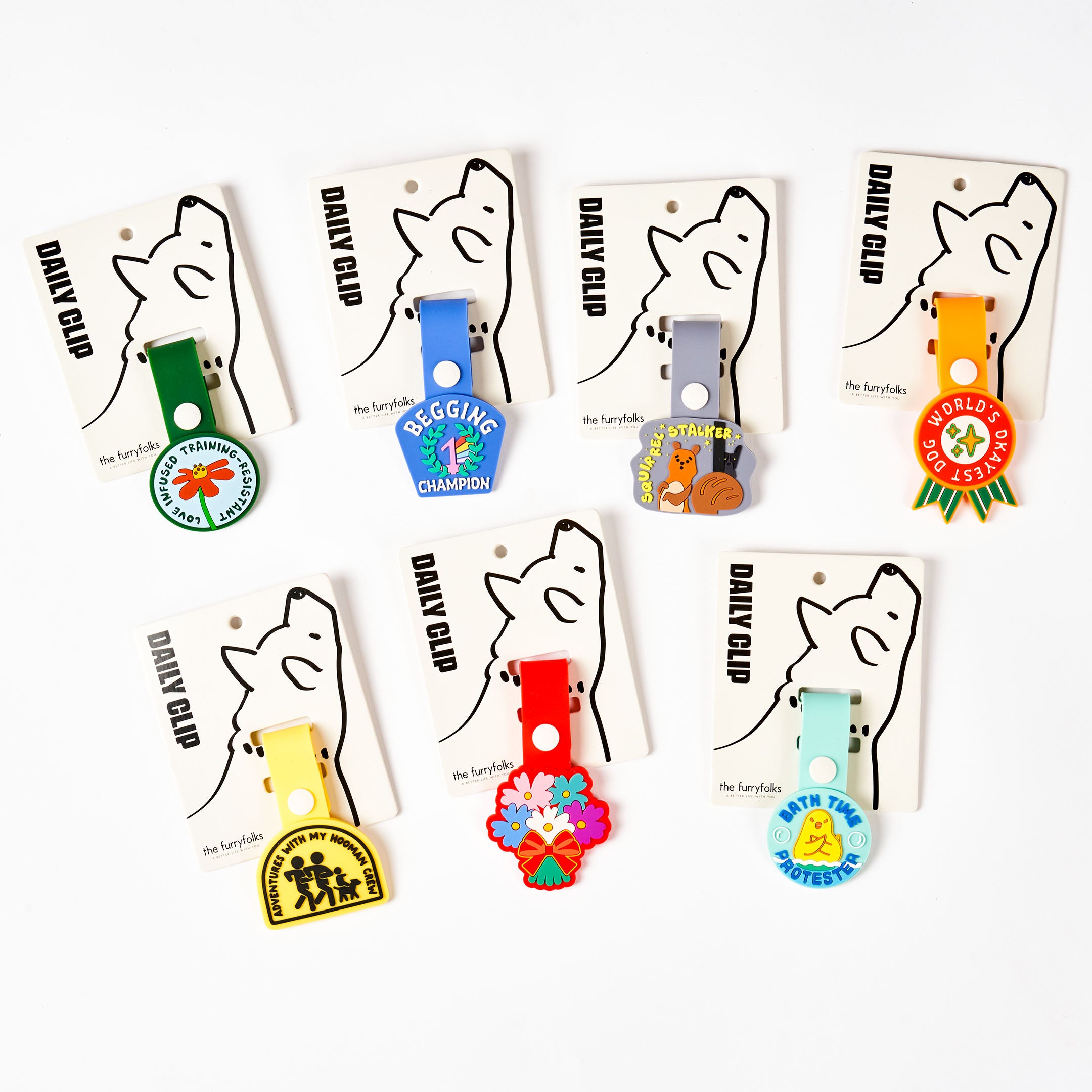 Seven colorful dog tags displayed on their packaging, each labeled ‘Daily Clip.’ The tags include designs such as ‘Love Infused Training-resistant,’ ‘Begging Champion,’ ‘Squirrel Stalker,’ ‘World’s Okayest Dog,’ ‘Adventures with My Hooman Crew,’ and ‘Bath Time Protester.’ Each tag is made of non-toxic rubber and features fun phrases and illustrations, showcasing the variety of available designs.