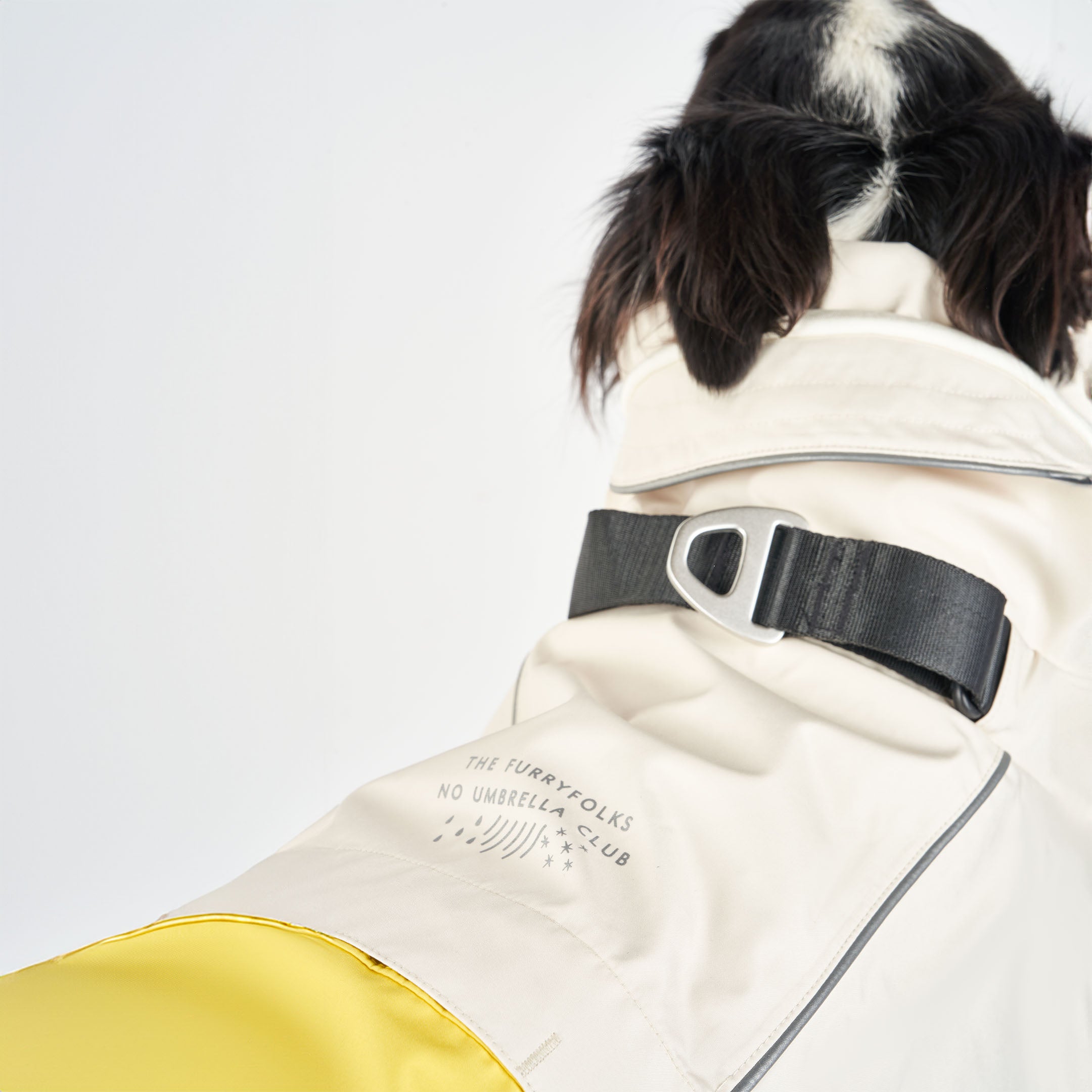 Close-up rear view of a large dog wearing a cream and yellow raincoat. The raincoat features a black buckle, reflective details, and the brand’s logo that reads “The Furryfolks - No Umbrella Club” on the back.