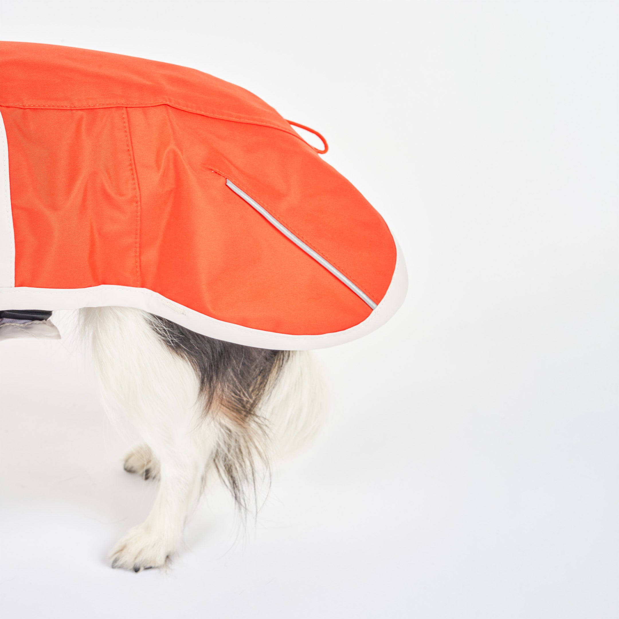 Side view of a small dog wearing an orange and gray raincoat with reflective accents. The raincoat features a large flap covering the back, offering protection from the rain and wind.