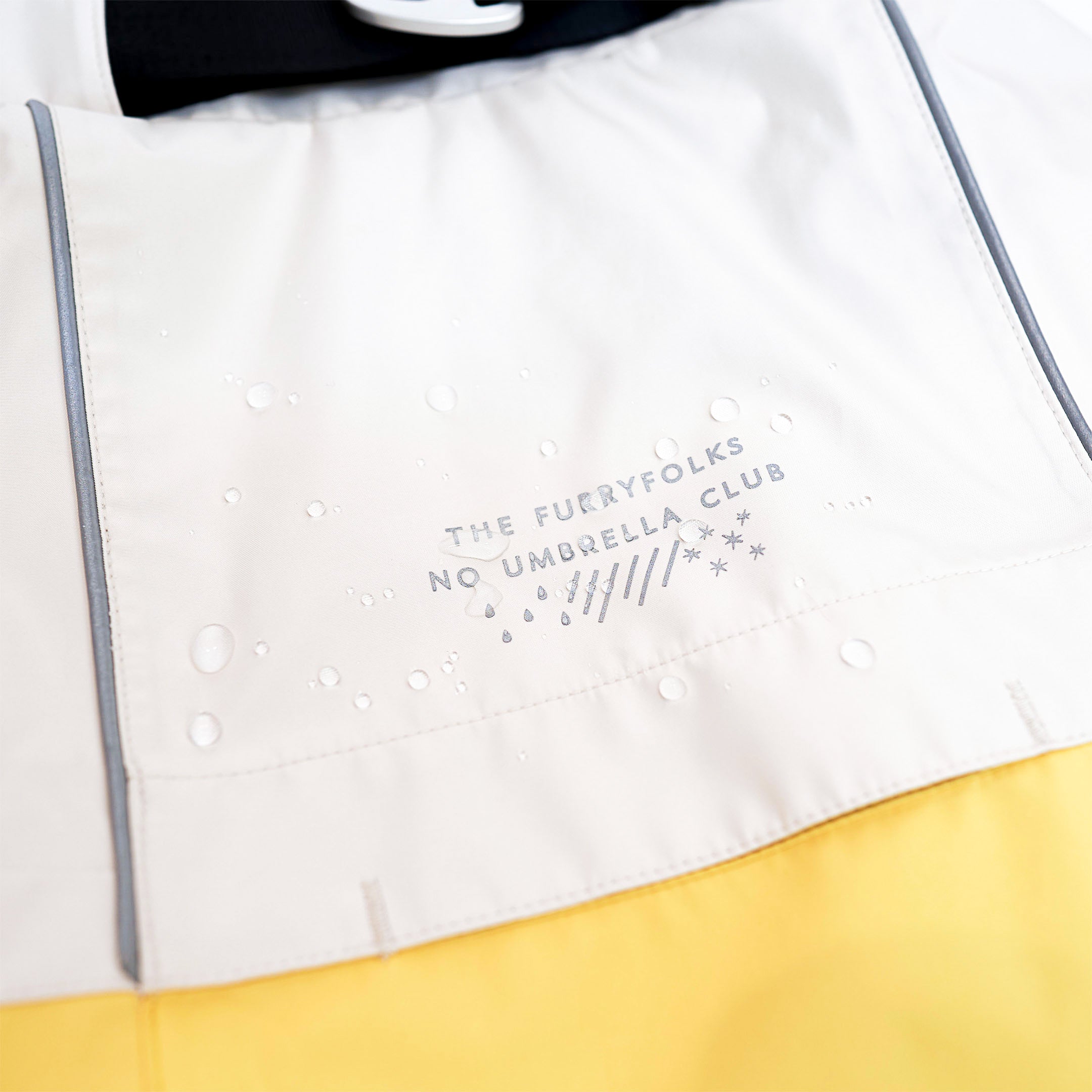 Close-up of a cream and yellow raincoat with water droplets on the back, showcasing its water-resistant material. The raincoat features a logo that reads “The Furryfolks - No Umbrella Club” and reflective accents for visibility.